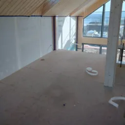 Wnętrze poddasza w trakcie wykańczania: surowe drewniane belki stropowe, ściany przygotowane do gładziowania z niebieską taśmą malarską, podłoga z płyt OSB, duże okno z widokiem na krajobraz.