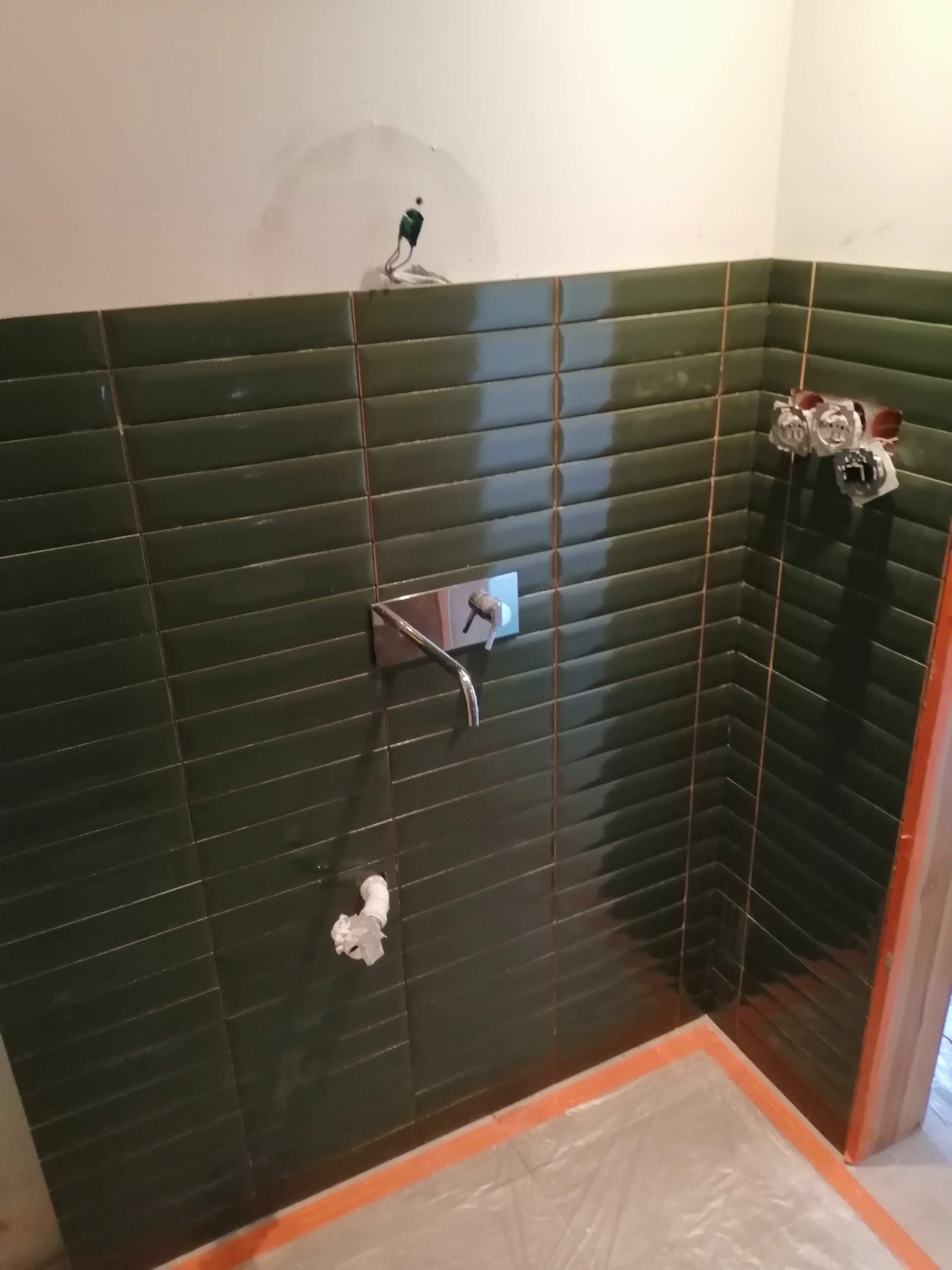 Ściana w łazience wyłożona ciemnozielonymi, podłużnymi płytkami z widoczną armaturą i instalacją elektryczną, podłoga zabezpieczona folią ochronną.