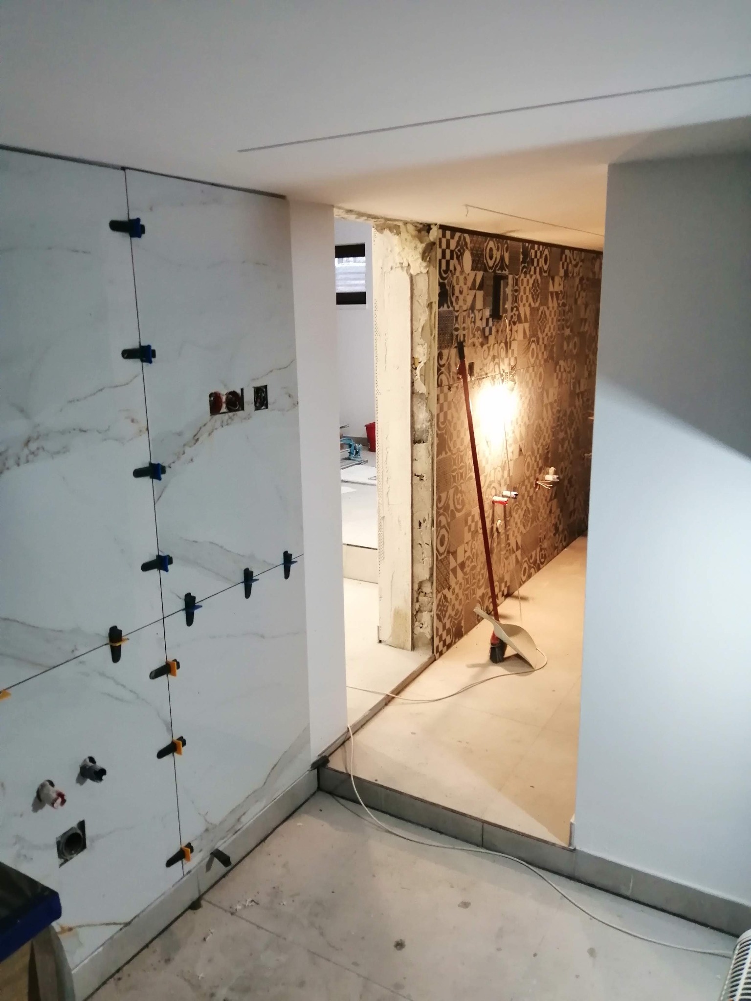 Ściana łazienki z białymi płytkami imitującymi marmur, zamocowanymi za pomocą klipsów poziomujących, widoczne przygotowane instalacje wodne i elektryczne, w tle fragment ściany z wzorzystymi...