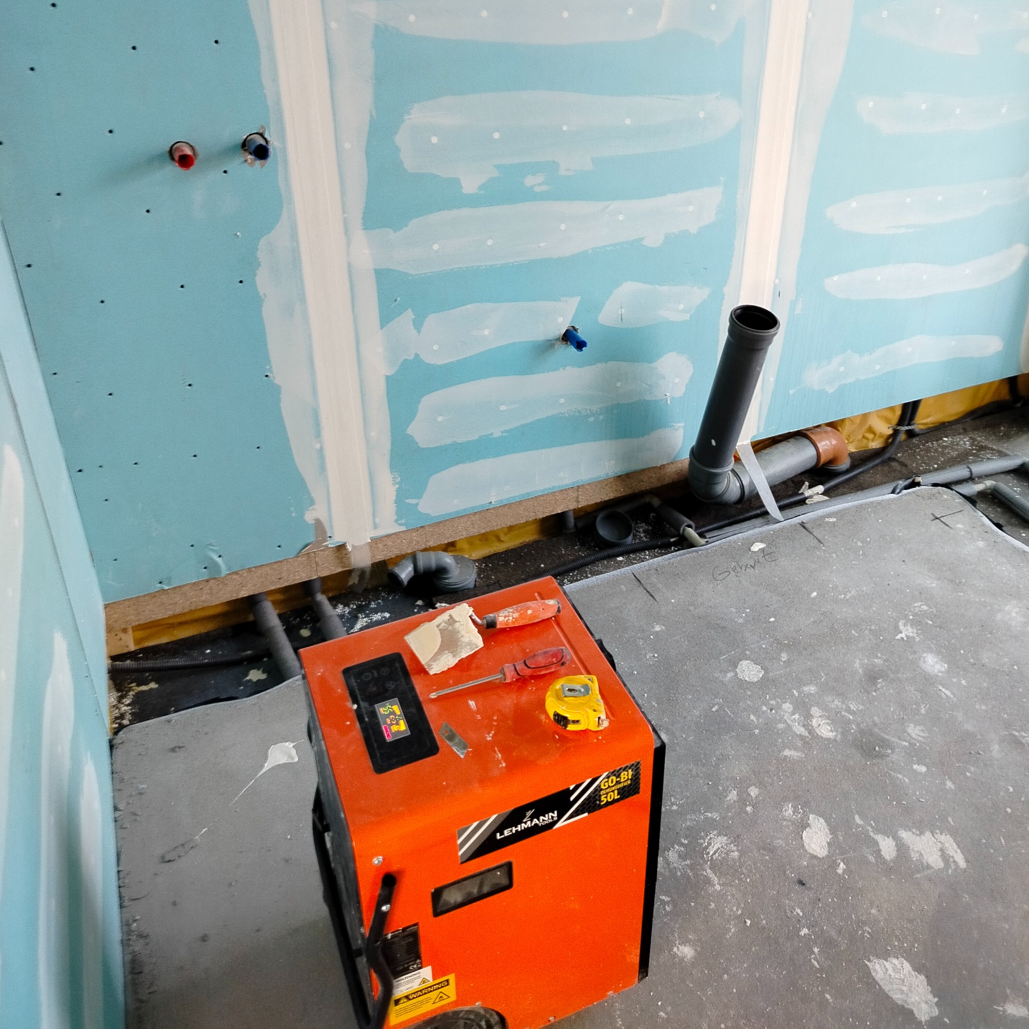Niedokończona łazienka z niebieskimi płytami gipsowo-kartonowymi, częściowo pokrytymi gładzią, z widocznymi rurami i pomarańczowym osuszaczem Lehmann 50L z narzędziami na górze.