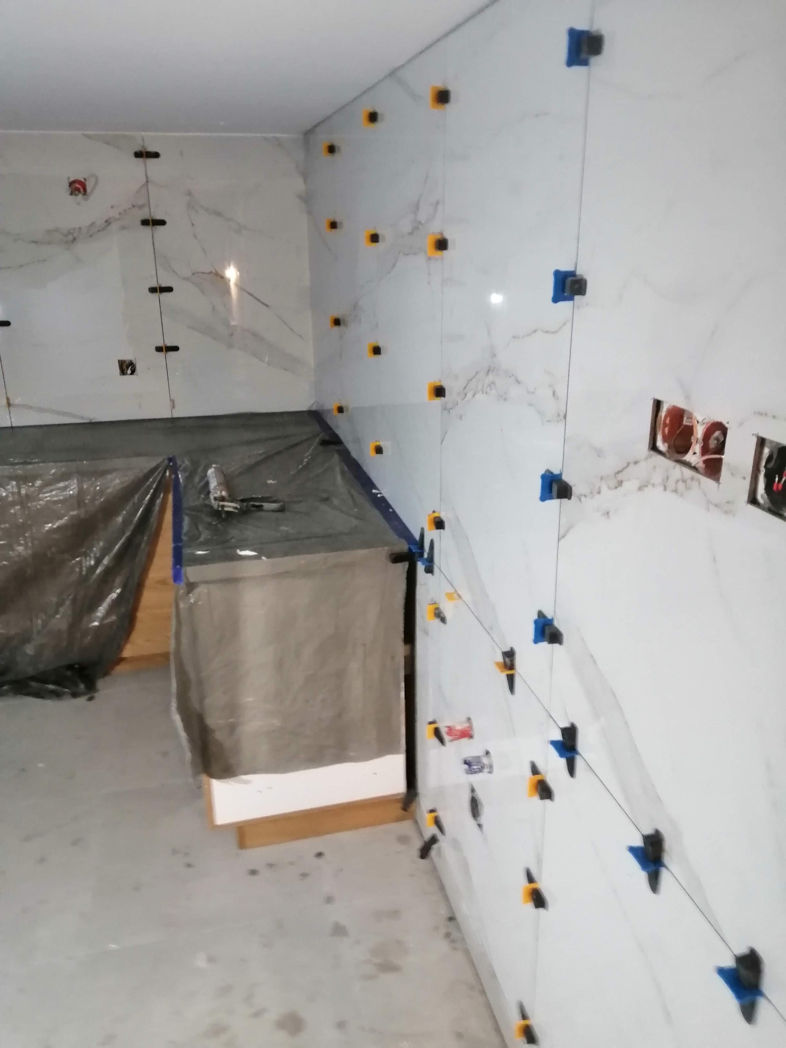 Ściana w trakcie wykańczania z białymi, marmurkowymi płytkami, użyte systemy poziomowania płytek (klipsy i kliny) w kolorach żółtym i niebieskim, widoczne puszki elektryczne i zabezpieczona folią...