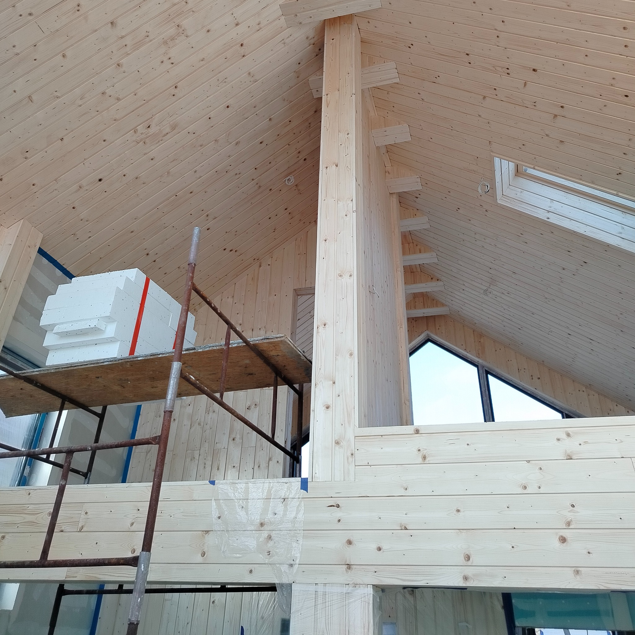 Wnętrze domu w trakcie budowy, widoczne drewniane ściany, strop i konstrukcja nośna, na rusztowaniu leżą białe płyty izolacyjne z czerwoną taśmą.