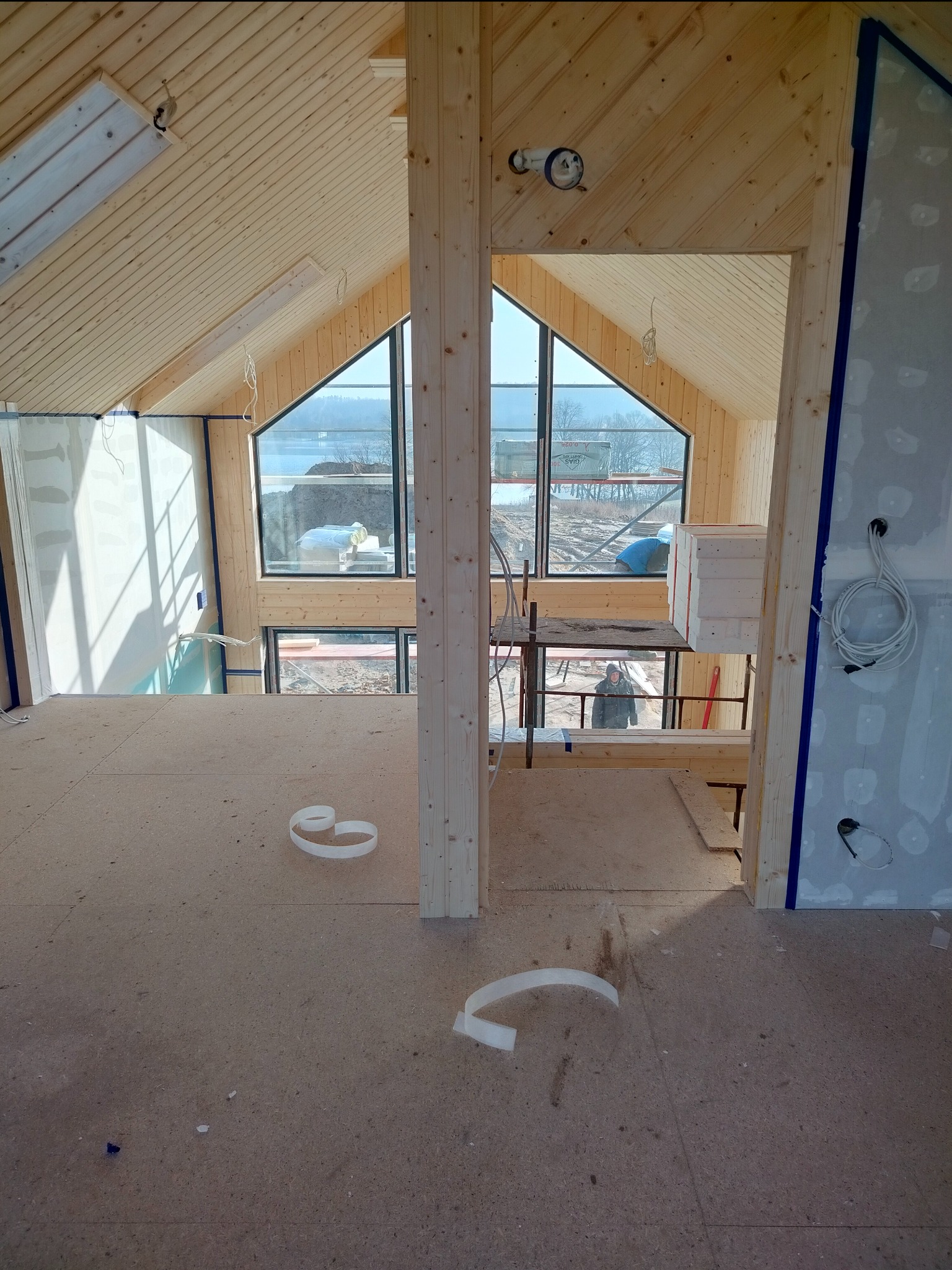 Wnętrze domu w trakcie wykańczania, widoczne drewniane panele na suficie, duże okna z widokiem na jezioro, ściany przygotowane do gładzi, podłoga z płyt OSB i resztki taśmy malarskiej.