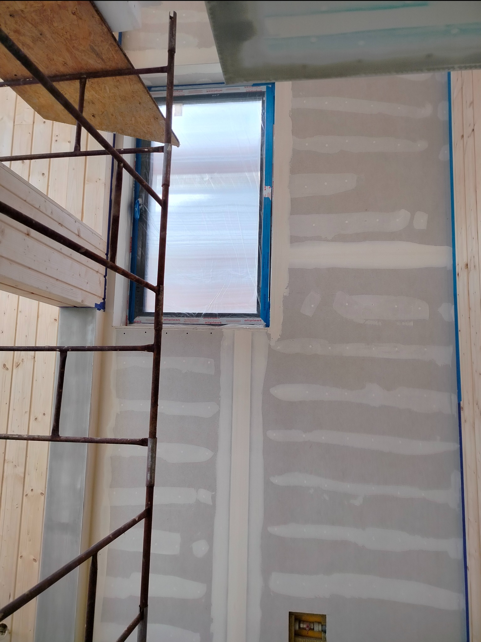 Ściana w trakcie prac wykończeniowych z widocznymi pasami nałożonej gładzi szpachlowej, okno zabezpieczone folią, rusztowanie w pomieszczeniu, ściany wykończone boazerią.