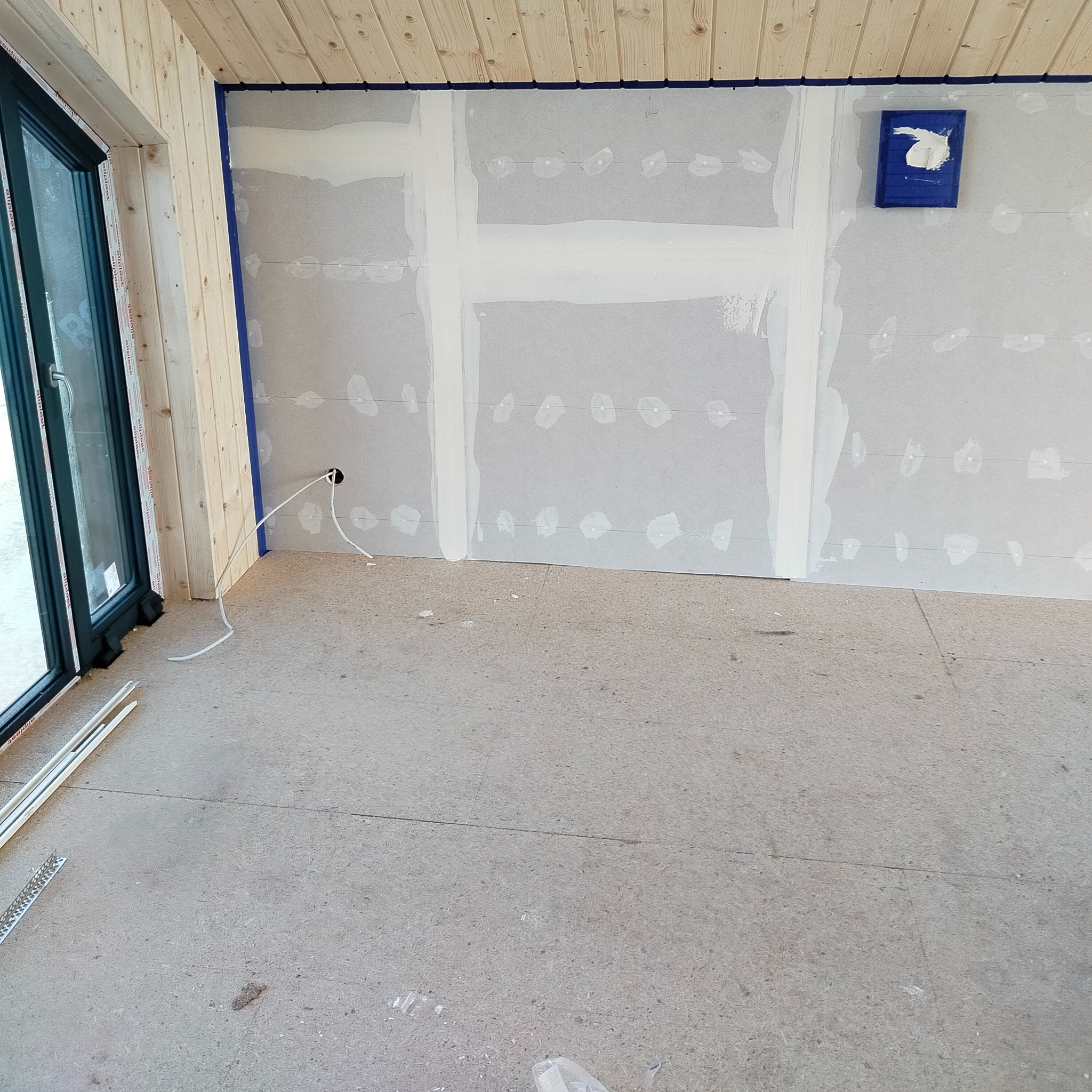 Wnętrze w trakcie prac wykończeniowych: zagruntowane płyty gipsowo-kartonowe na ścianach z widocznymi łączeniami wypełnionymi gładzią, zabezpieczone niebieską taśmą malarską, drewniana boazeria...