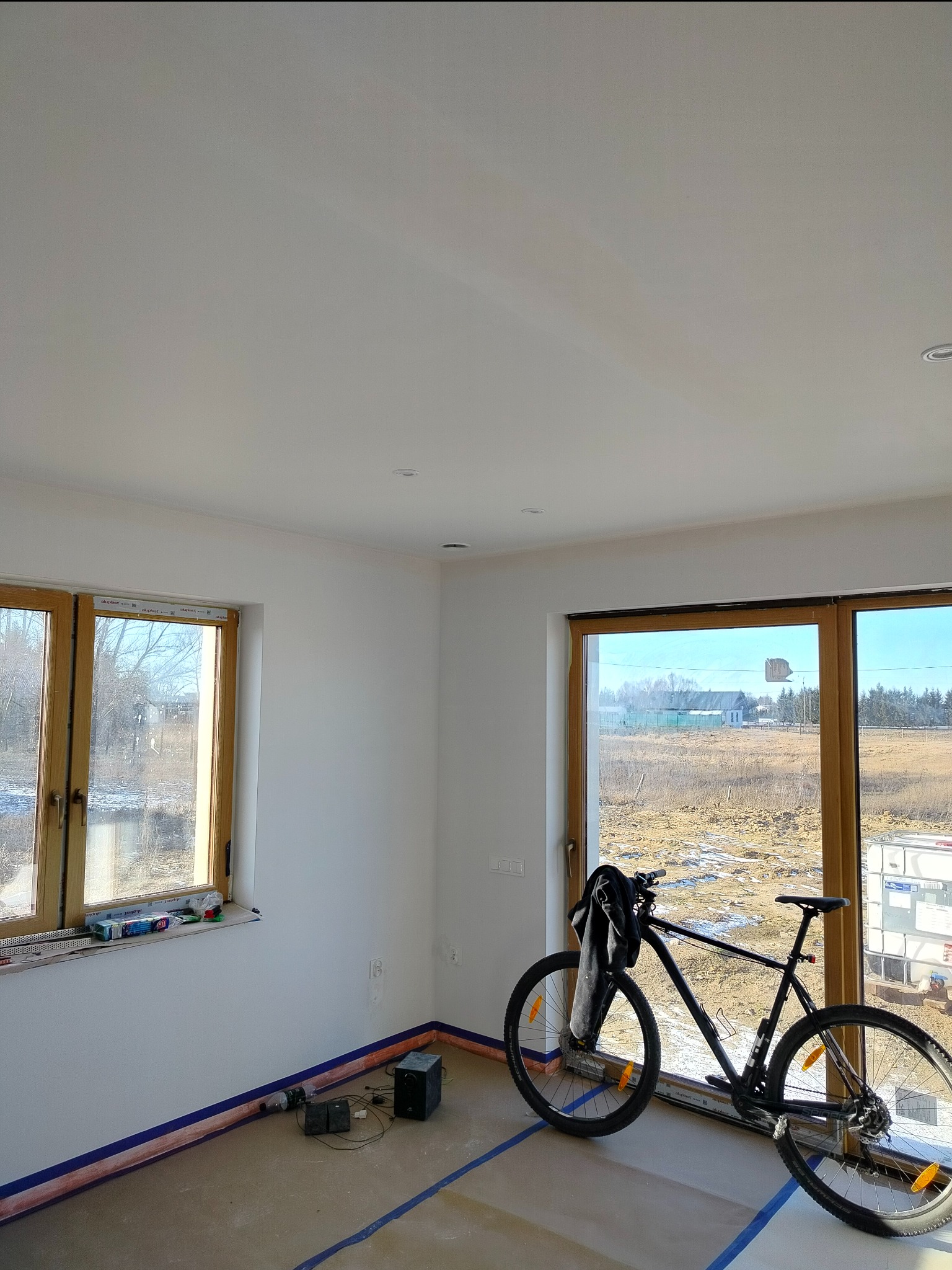 Wnętrze pokoju w trakcie remontu, z zabezpieczoną podłogą taśmą malarską, świeżo pomalowane ściany, rower oparty o okno oraz widok na zewnątrz.