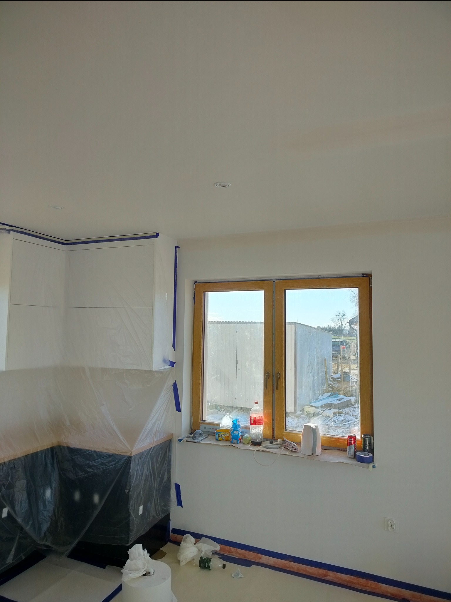 Przygotowanie kuchni do malowania: meble zabezpieczone folią, okna z drewnianą ramą oklejone taśmą malarską, widoczne butelki i puszki na parapecie.