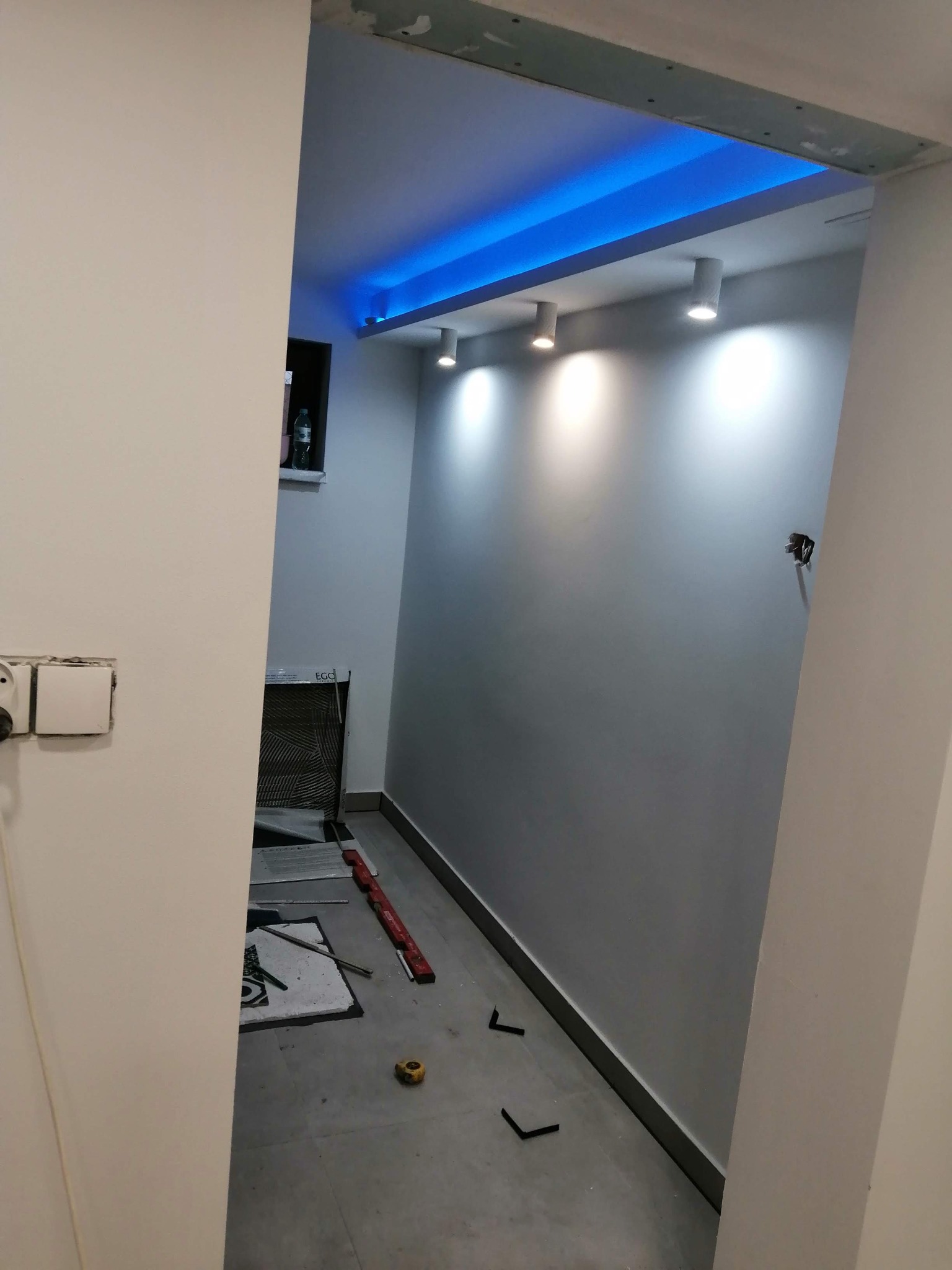 Wnętrze w trakcie prac wykończeniowych z podświetlonym sufitem, widoczne płytki podłogowe, poziomica i lampy punktowe.