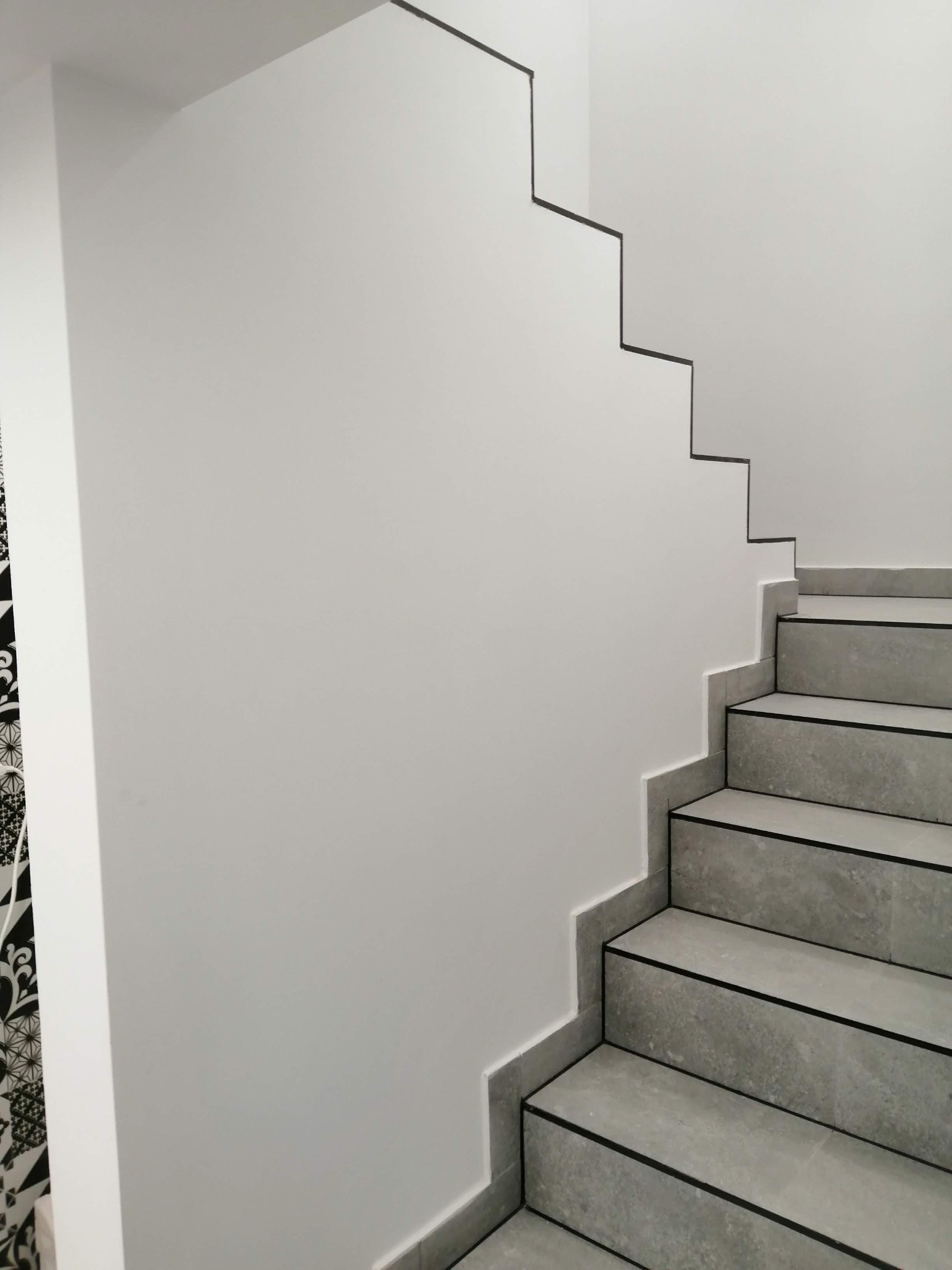 Minimalistyczne schody z szarych płytek z czarnymi akcentami, przylegające do białej ściany z dekoracyjnym wykończeniem, kontrastujące z czarno-białą tapetą w tle.
