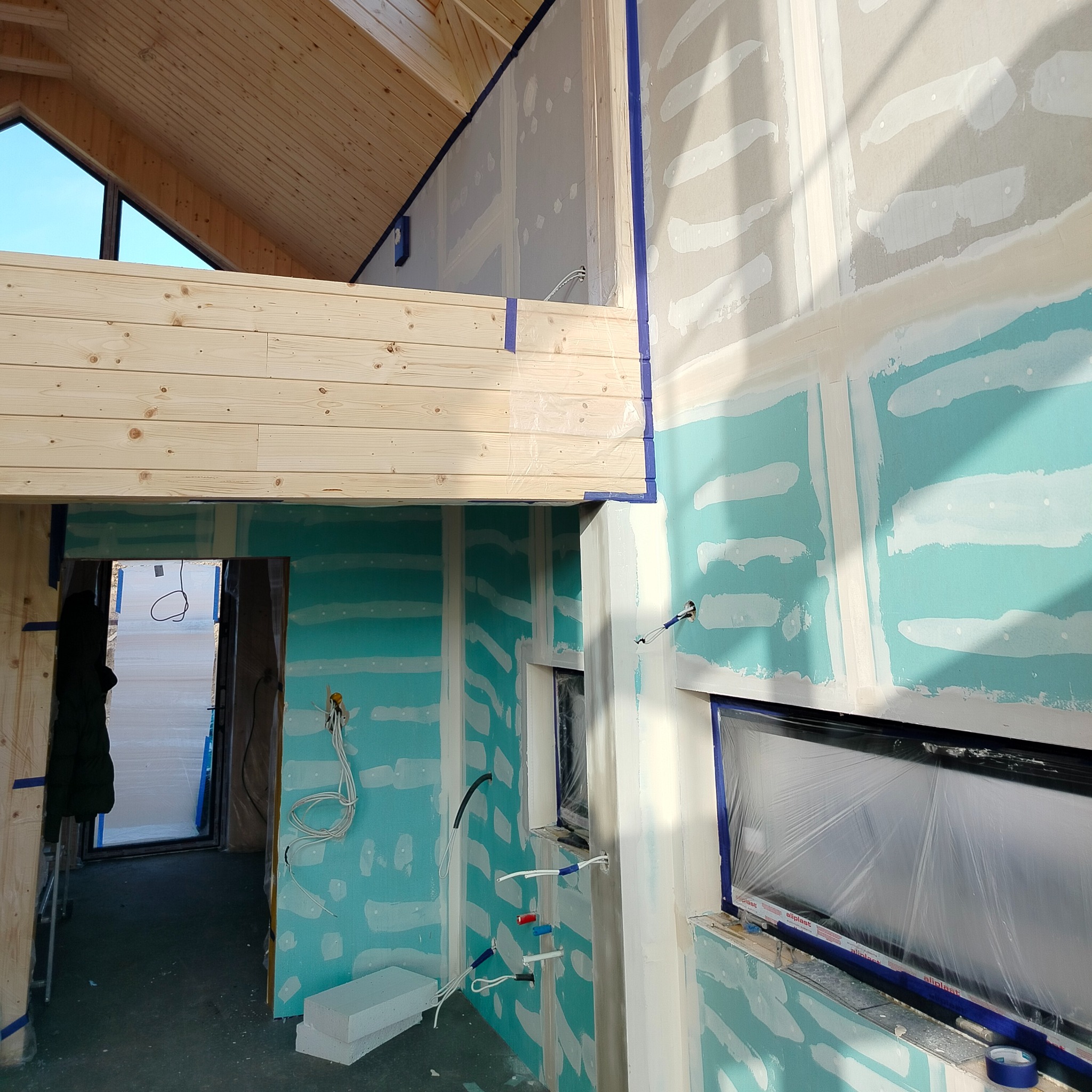 Wnętrze w trakcie wykańczania: surowa konstrukcja drewniana stropu, ściany z płyt gipsowo-kartonowych z widocznymi łączeniami i taśmą malarską, okna zabezpieczone folią ochronną, widoczne przewody...