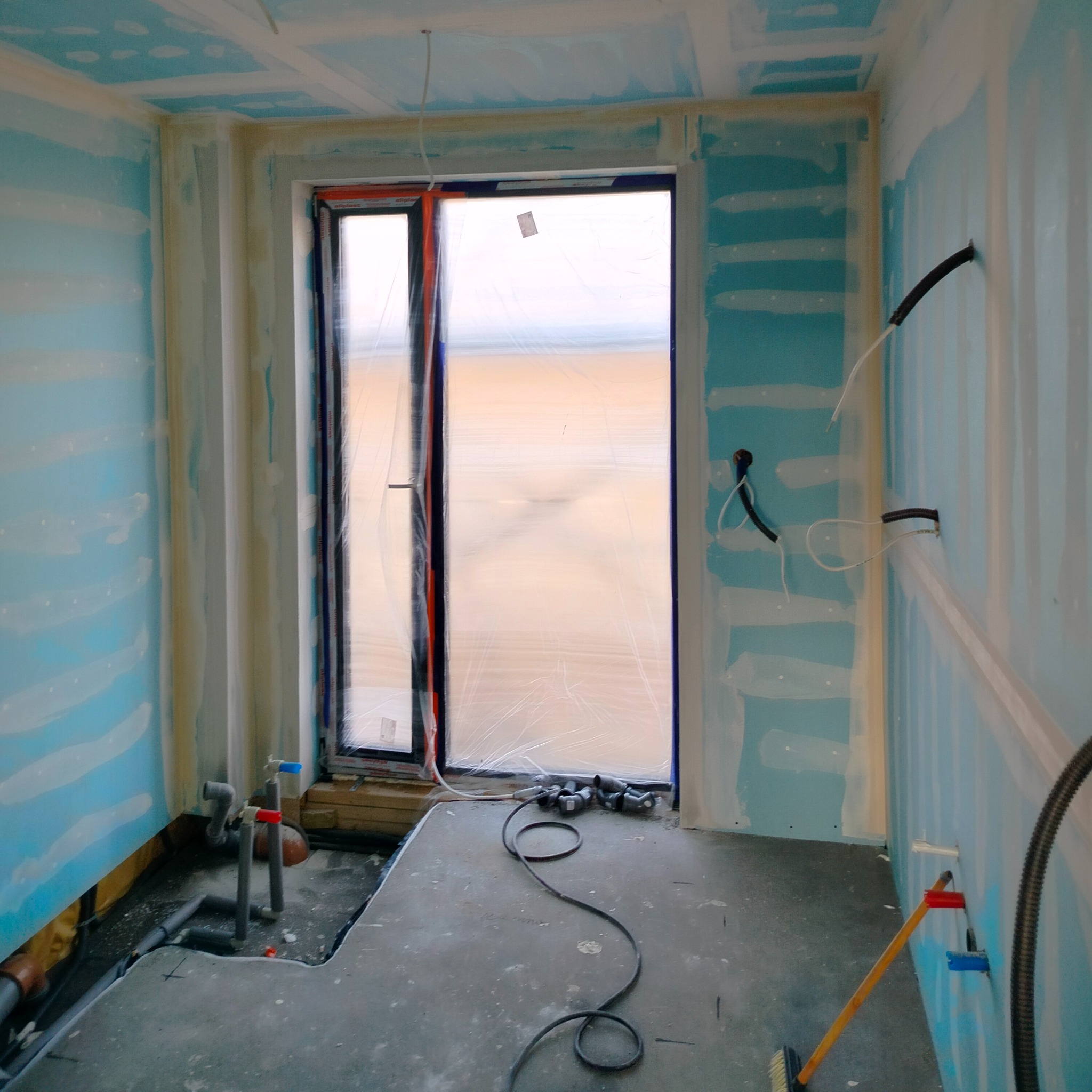 Wnętrze w trakcie remontu: ściany z płyt gipsowo-kartonowych, częściowo zagruntowane, okno zabezpieczone folią, widoczne elementy instalacji hydraulicznej i elektrycznej, na podłodze narzędzia...