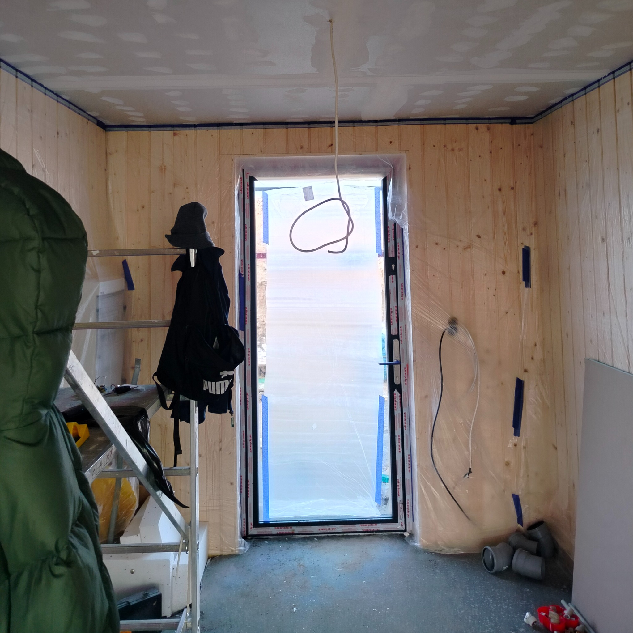 Wnętrze w trakcie prac wykończeniowych: ściany obłożone boazerią zabezpieczone folią, sufit po wstępnym szpachlowaniu, oklejone taśmą malarską. Na podłodze rury kanalizacyjne, a obok drabina...