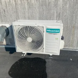 Biała jednostka zewnętrzna klimatyzacji Hisense na dachu, widoczny fragment instalacji wentylacyjnej. Teksturowana ściana w tle. Widoczny cień osoby.