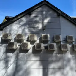 Elewacja budynku z zainstalowanymi jednostkami zewnętrznymi klimatyzacji Daikin, cień drzewa na ścianie. Błękitne niebo w tle. Liczne klimatyzatory.