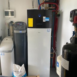 DS Klimatyzacje Daniel Spychała - Pompa ciepła Daikin w kotłowni, obok zmiękczacz wody i filtr, widoczne rury instalacji grzewczej, worek z solą i karton. W tle białe ściany i instalacje.