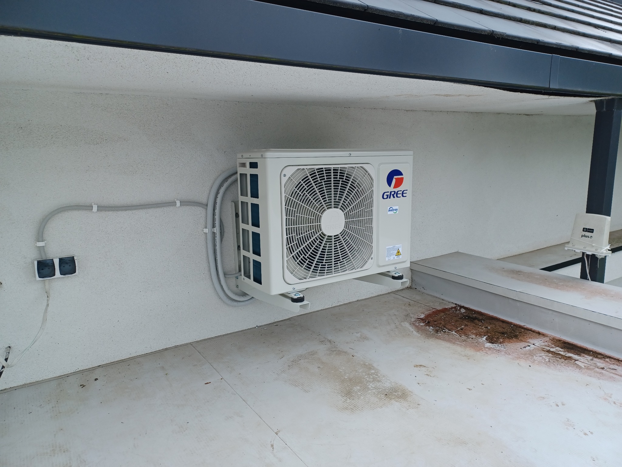 Zewnętrzna jednostka klimatyzatora marki Gree zamontowana na wspornikach przy ścianie budynku, z widocznymi przewodami w peszlach i gniazdkami elektrycznymi.