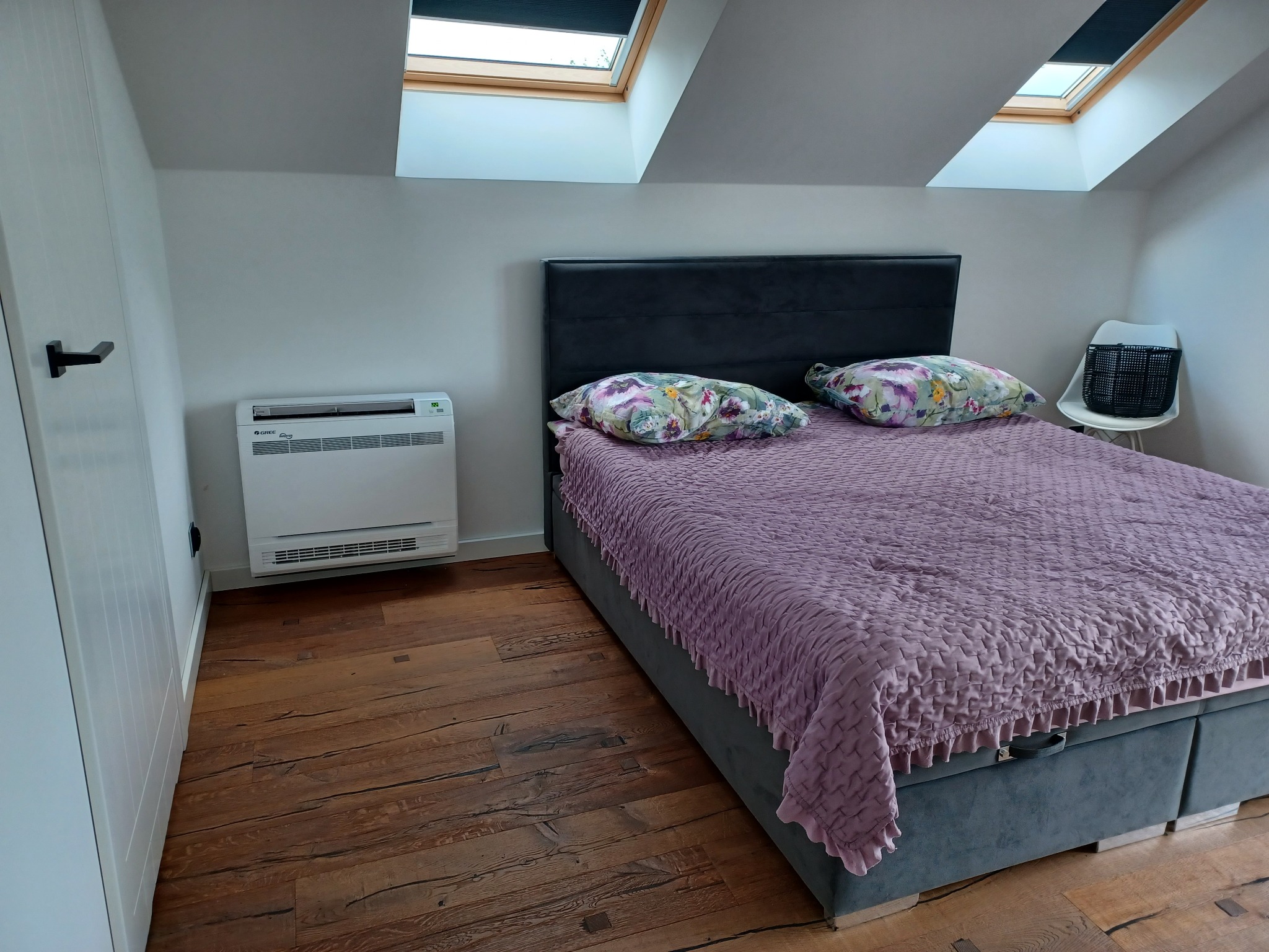 Biały klimatyzator podłogowy firmy Gree zamontowany w sypialni na poddaszu z drewnianą podłogą i oknami dachowymi.