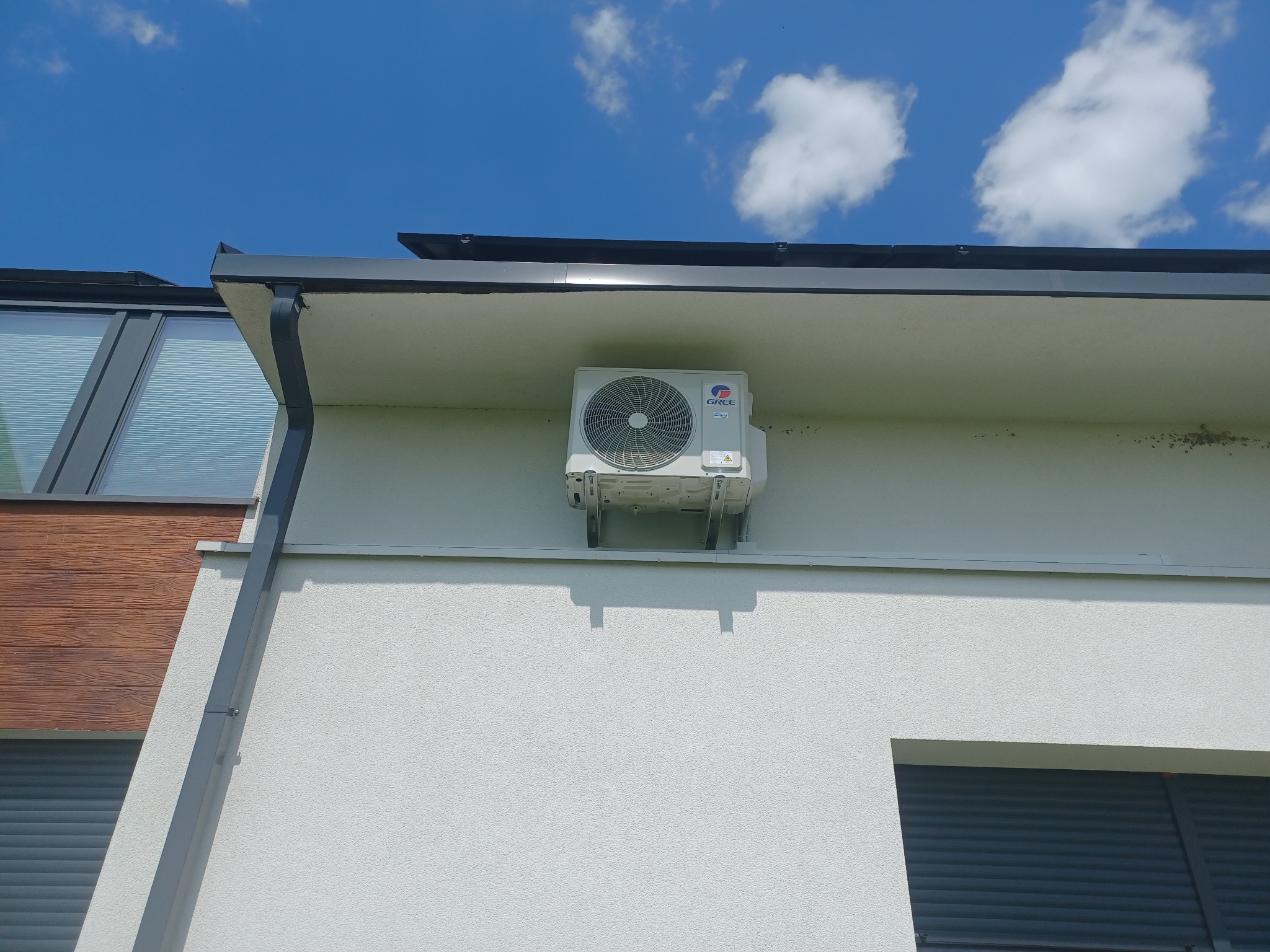 Zewnętrzna jednostka klimatyzacji marki Gree zamontowana na elewacji jasnego budynku z widocznymi oknami i rynną na tle błękitnego nieba z chmurami.