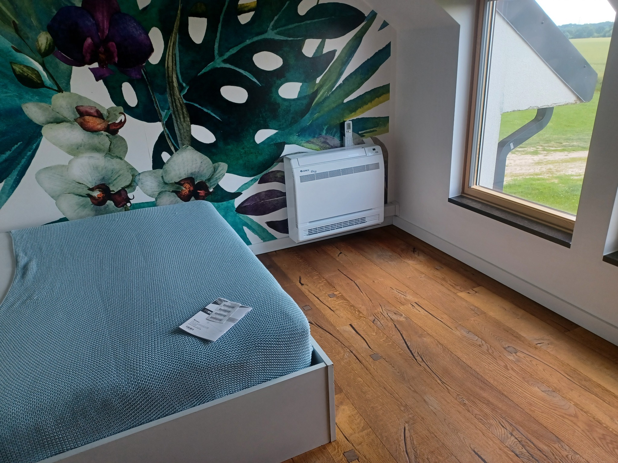 Podłogowy klimatyzator Gree w sypialni w Szczecinie, z dekoracyjną tapetą w motywy roślinne i widokiem na zewnątrz przez okno.
