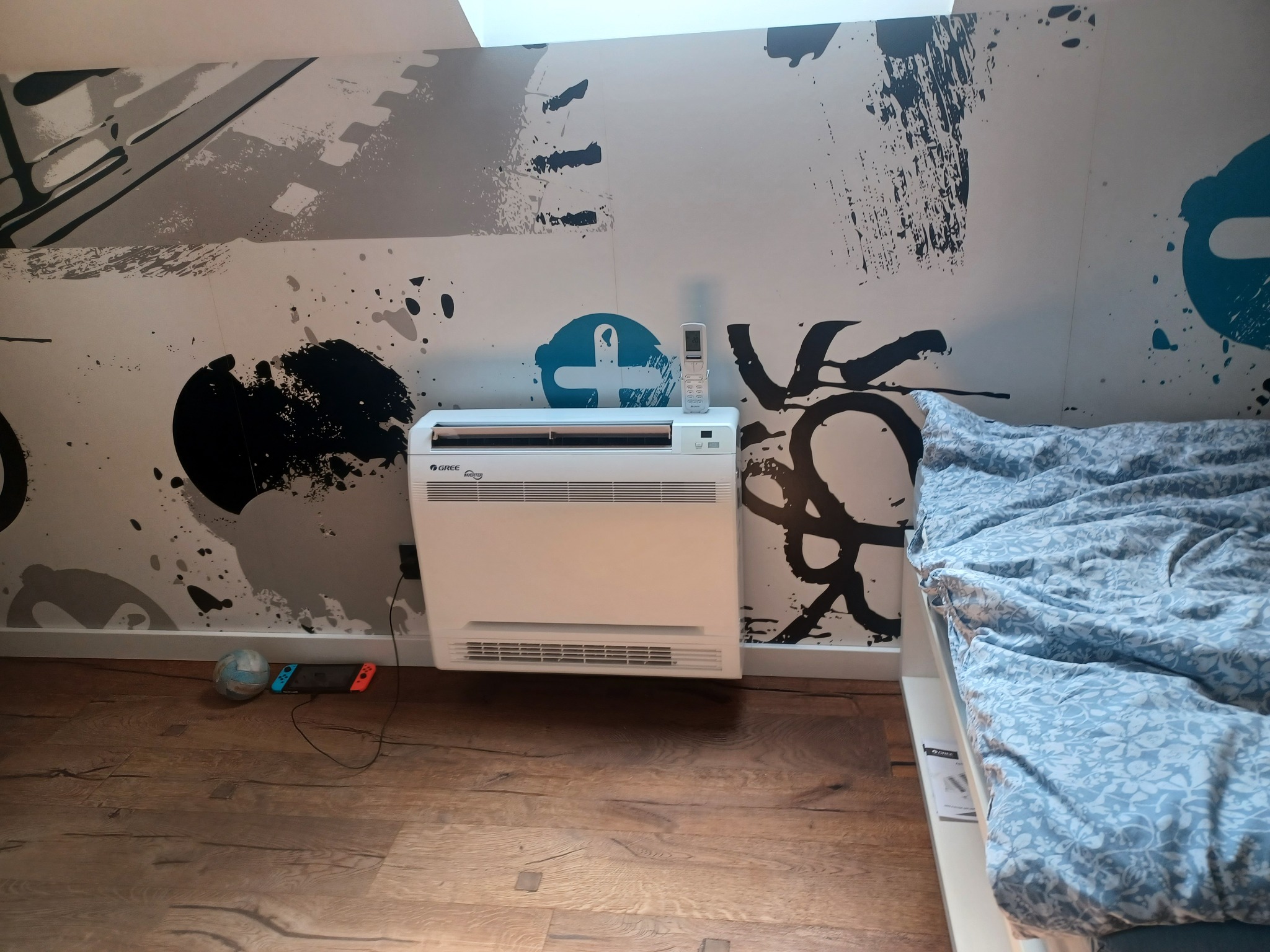 Biały klimatyzator przypodłogowy marki Gree w pokoju z designerską ścianą w stylu graffiti, obok łóżka z niebieską pościelą i konsoli do gier na drewnianej podłodze.
