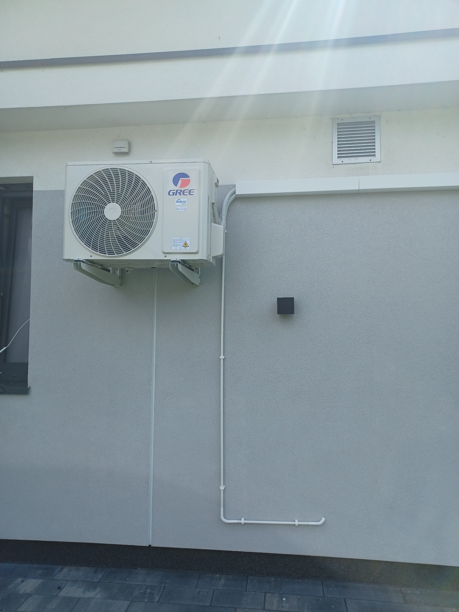 Biała jednostka zewnętrzna klimatyzatora marki Gree zamontowana na szarej, otynkowanej ścianie budynku z widocznymi przewodami instalacyjnymi i kratką wentylacyjną.