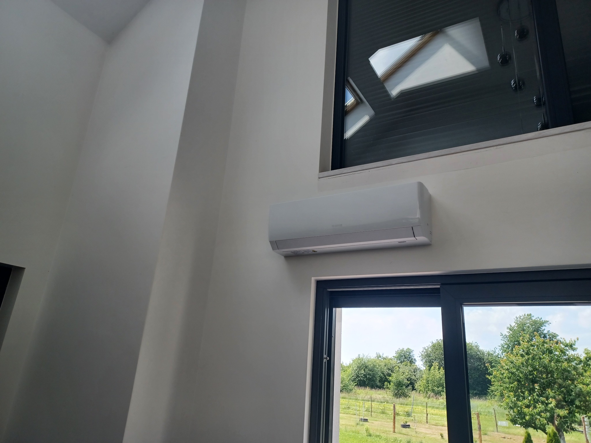 Biały klimatyzator ścienny zamontowany nad oknem z widokiem na zieleń, w nowoczesnym wnętrzu z oknem dachowym.