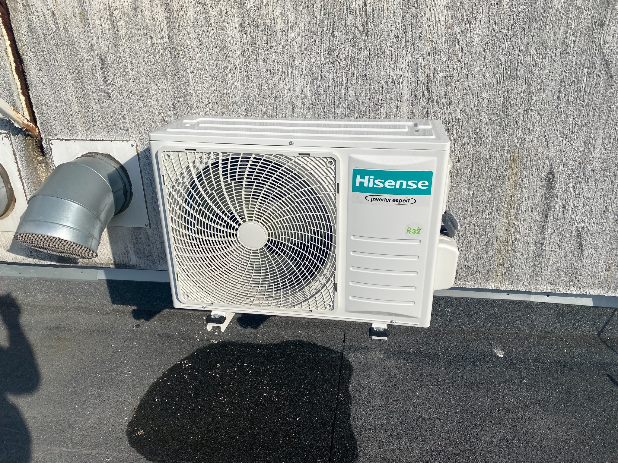 Biała jednostka zewnętrzna klimatyzacji Hisense na dachu, widoczny fragment instalacji wentylacyjnej. Teksturowana ściana w tle. Widoczny cień osoby.