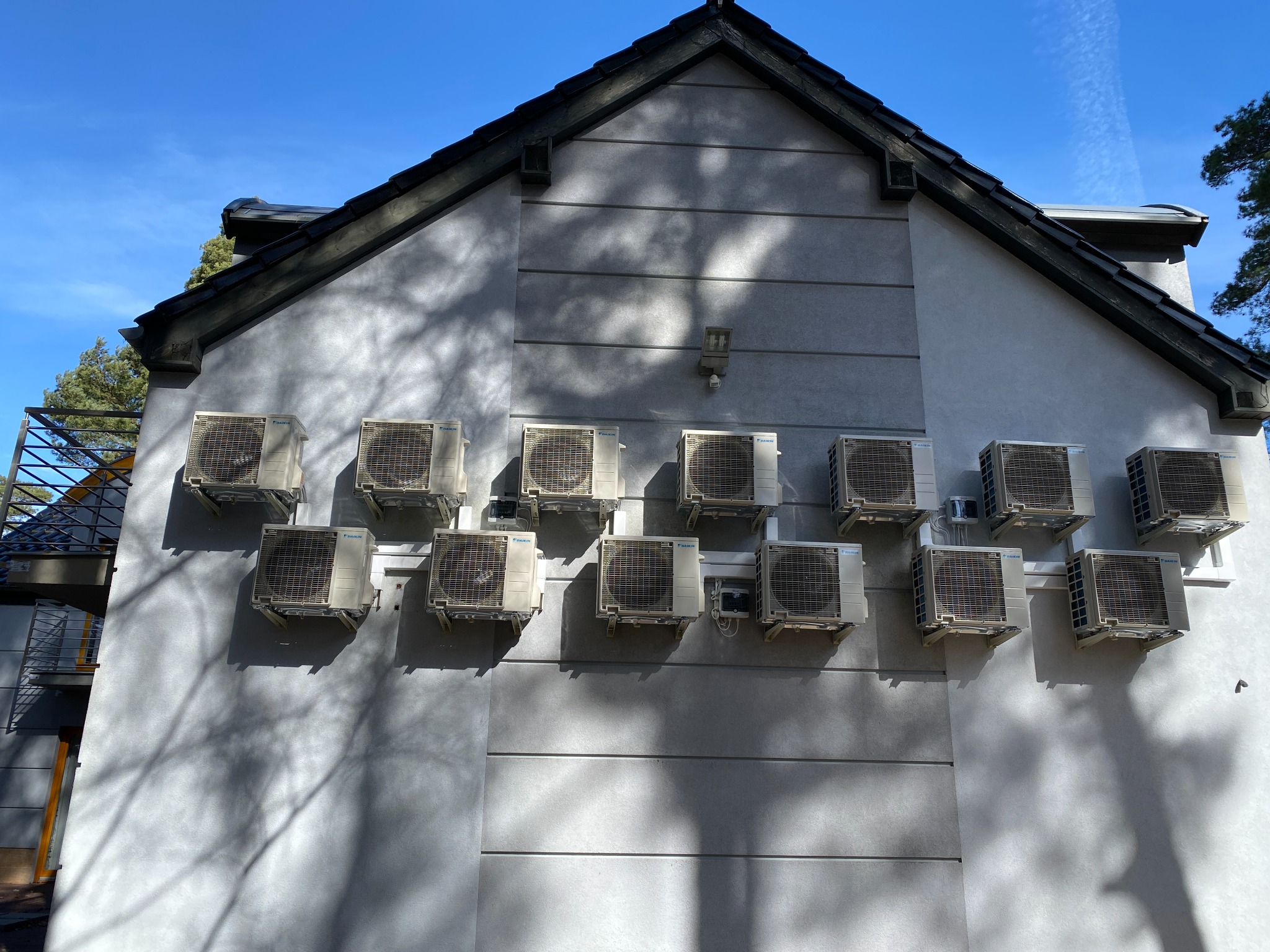 Elewacja budynku z zainstalowanymi jednostkami zewnętrznymi klimatyzacji Daikin, cień drzewa na ścianie. Błękitne niebo w tle. Liczne klimatyzatory.