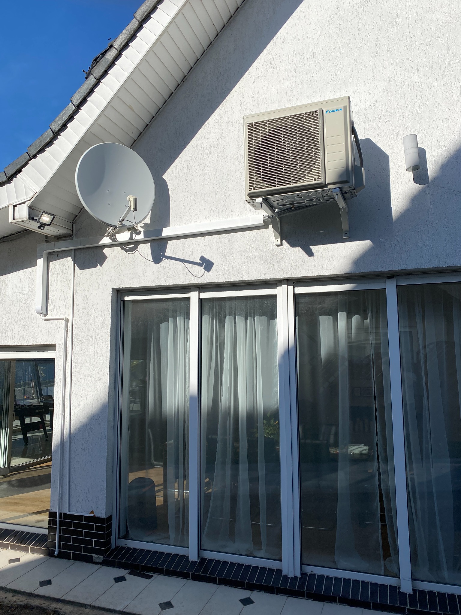 Biała elewacja domu z zamontowaną klimatyzacją Daikin, anteną satelitarną i lampą zewnętrzną. Widoczne przesuwne okna z zasłonami.