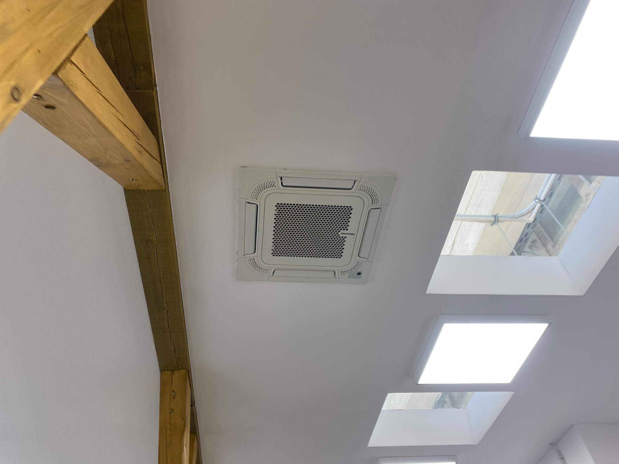 Kasetonowa jednostka klimatyzacji zamontowana na suficie w pomieszczeniu z drewnianą belką i oknami dachowymi. Widok z dołu.