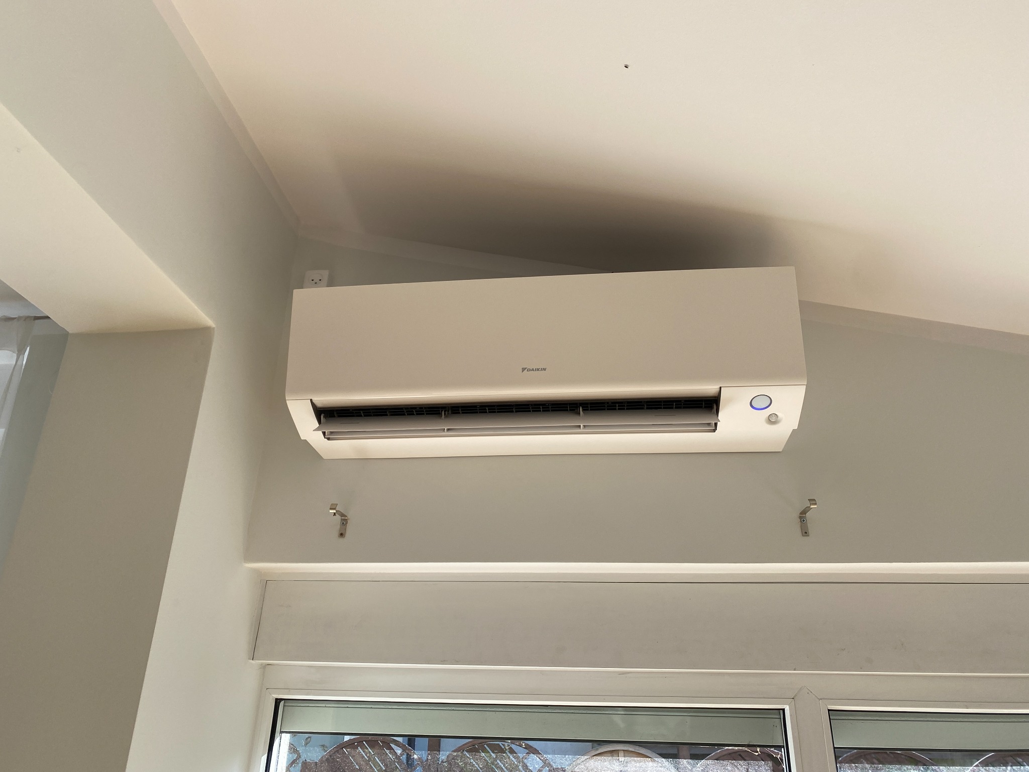 Biała klimatyzacja Daikin zamontowana na ścianie w jasnym pomieszczeniu z oknem. Widoczne haki montażowe i gniazdko elektryczne w górnym rogu.