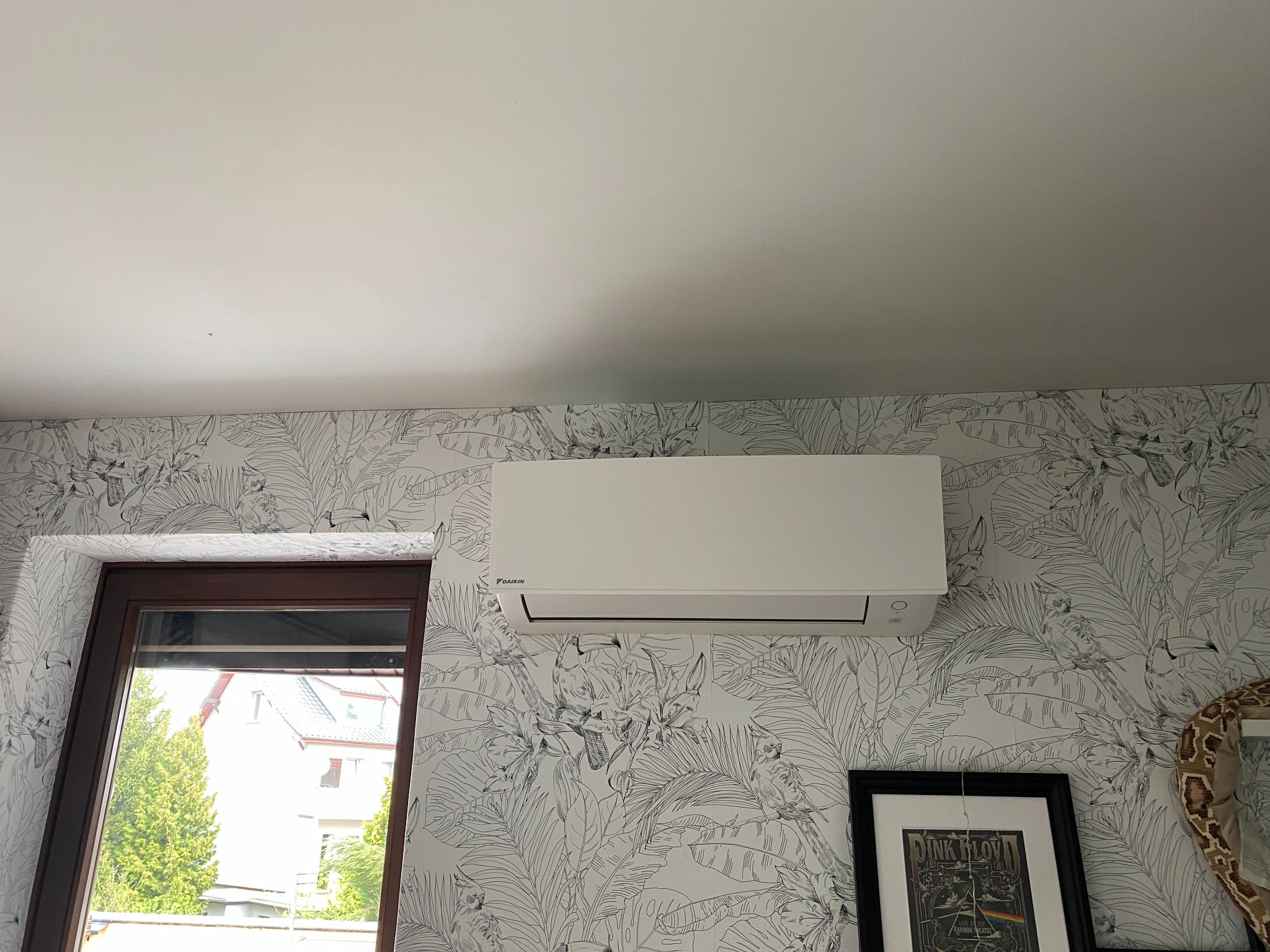 Biała klimatyzacja Daikin na ścianie z tropikalną tapetą. Obok okno i plakat Pink Floyd. Nowoczesny design, domowe wnętrze.