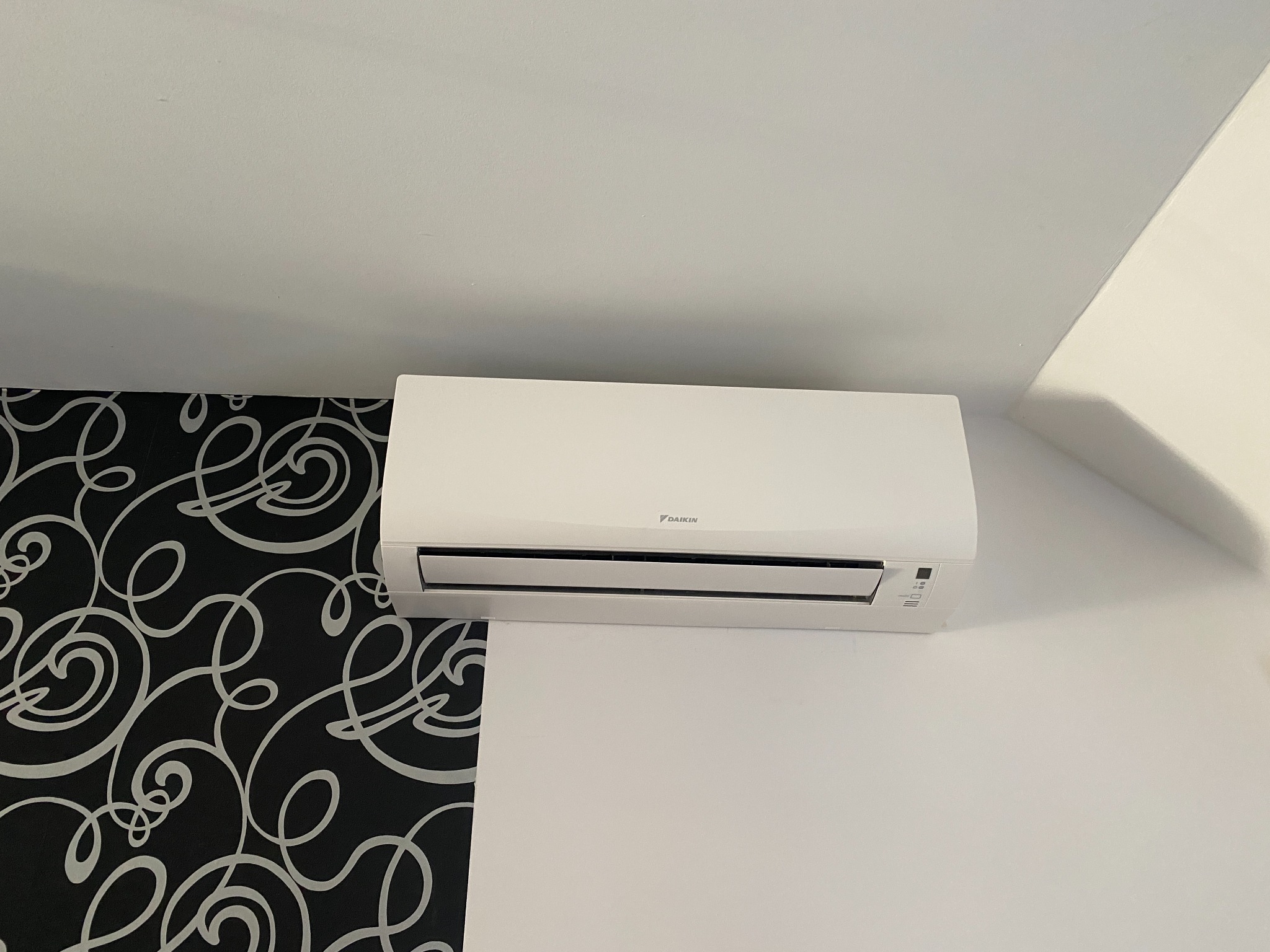 Biały klimatyzator Daikin na ścianie z wzorzystą tapetą. Urządzenie zamontowane w rogu pokoju, widoczne detale obudowy i panel sterowania.
