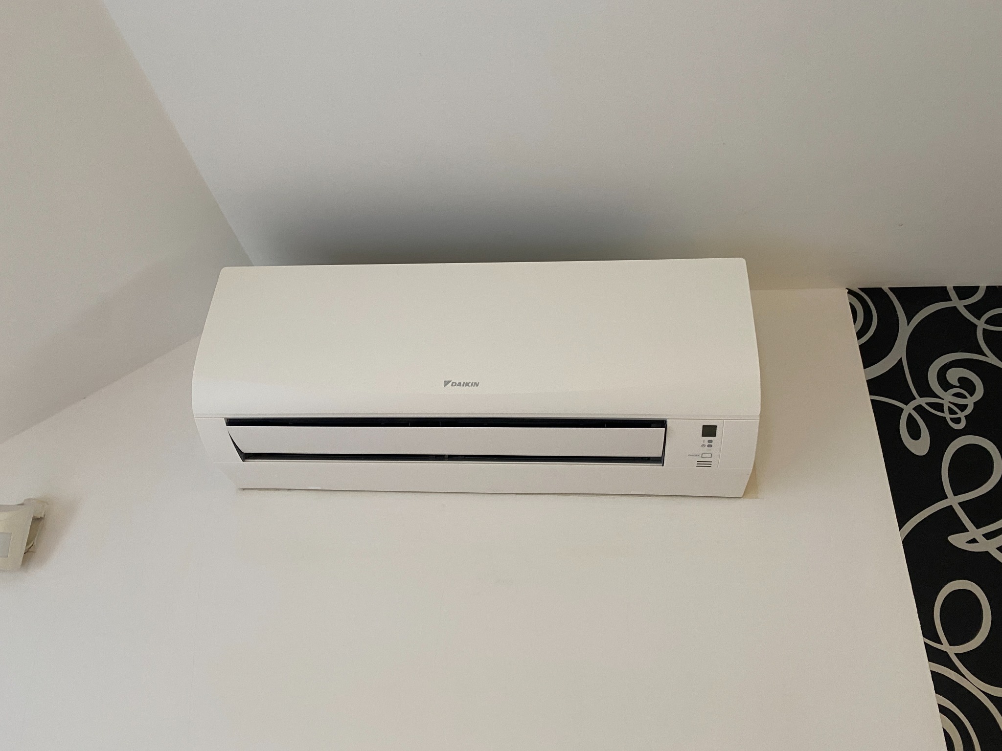Biała klimatyzacja Daikin zamontowana na ścianie w pomieszczeniu ze skośnym sufitem i dekoracyjną tapetą po prawej stronie.
