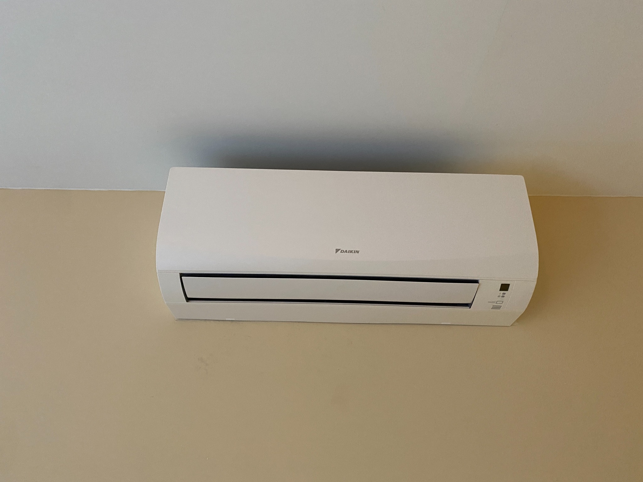 Biała klimatyzacja Daikin, zamontowana na beżowej ścianie, z bliska. Widoczne detale obudowy i panel sterowania. Minimalistyczny design.