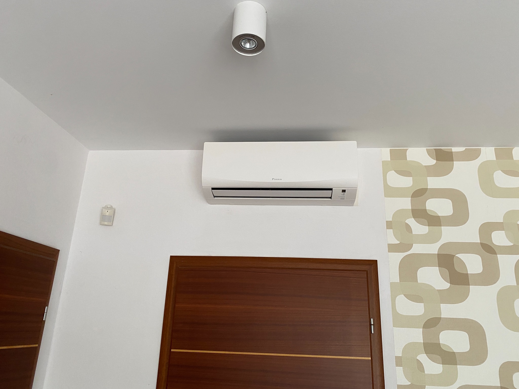 Biały klimatyzator Daikin na ścianie nad brązowymi drzwiami, obok wzorzystej tapety. Na suficie widoczna okrągła lampa.