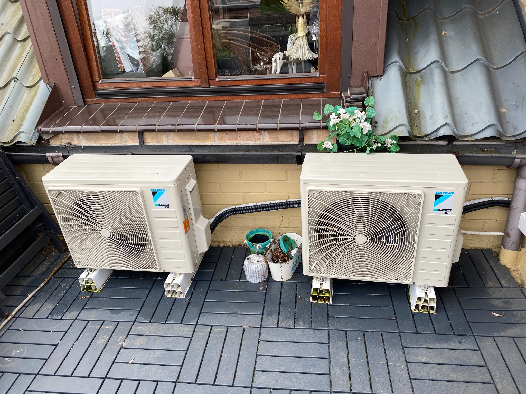 Dwa klimatyzatory Daikin na szarym tarasie, zamontowane na białych wspornikach, z doniczkami między nimi i oknem w tle.