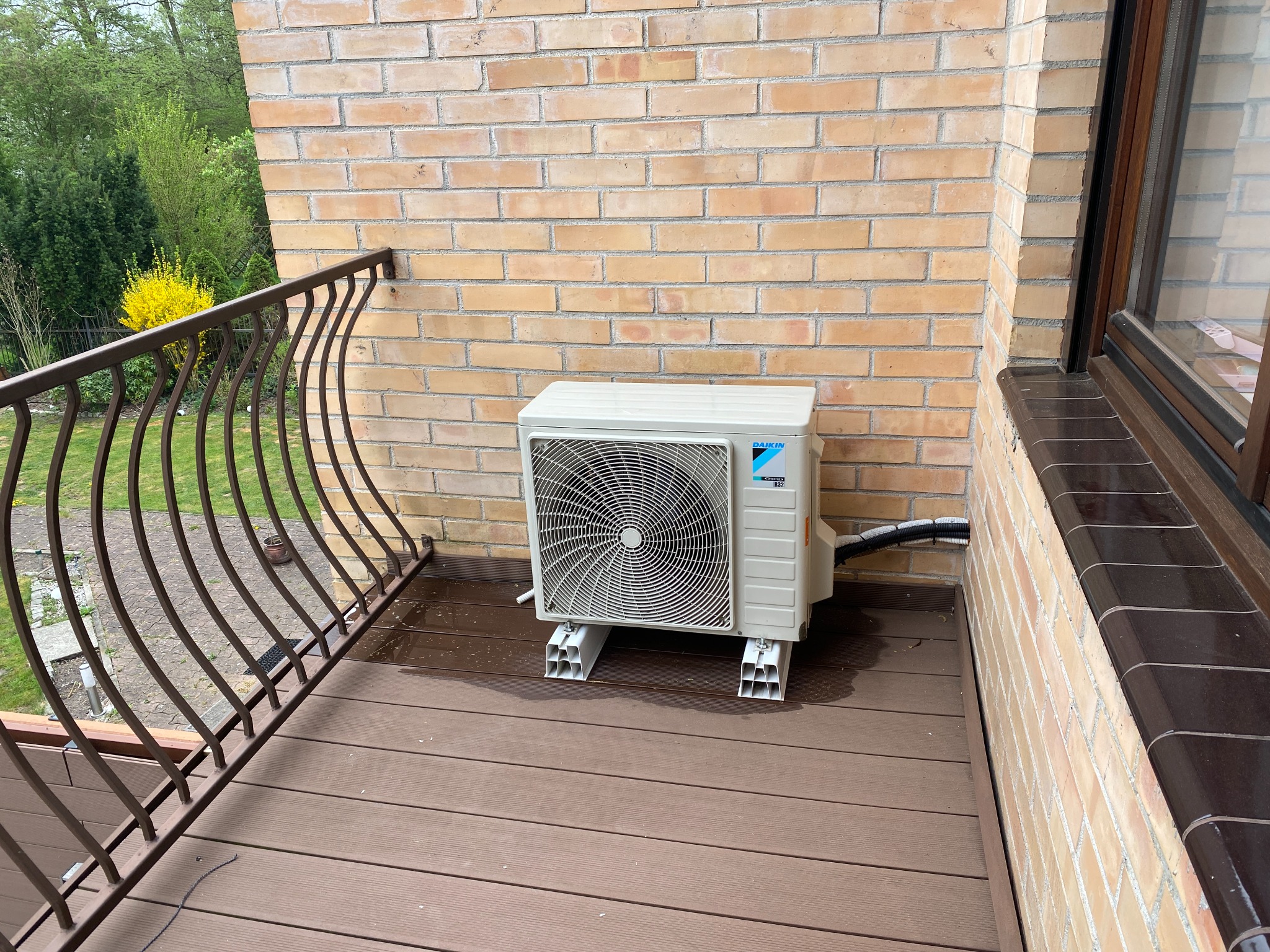 Biała jednostka zewnętrzna klimatyzacji Daikin na balkonie z brązową podłogą, zamontowana na wspornikach. W tle ceglana ściana i zieleń drzew.