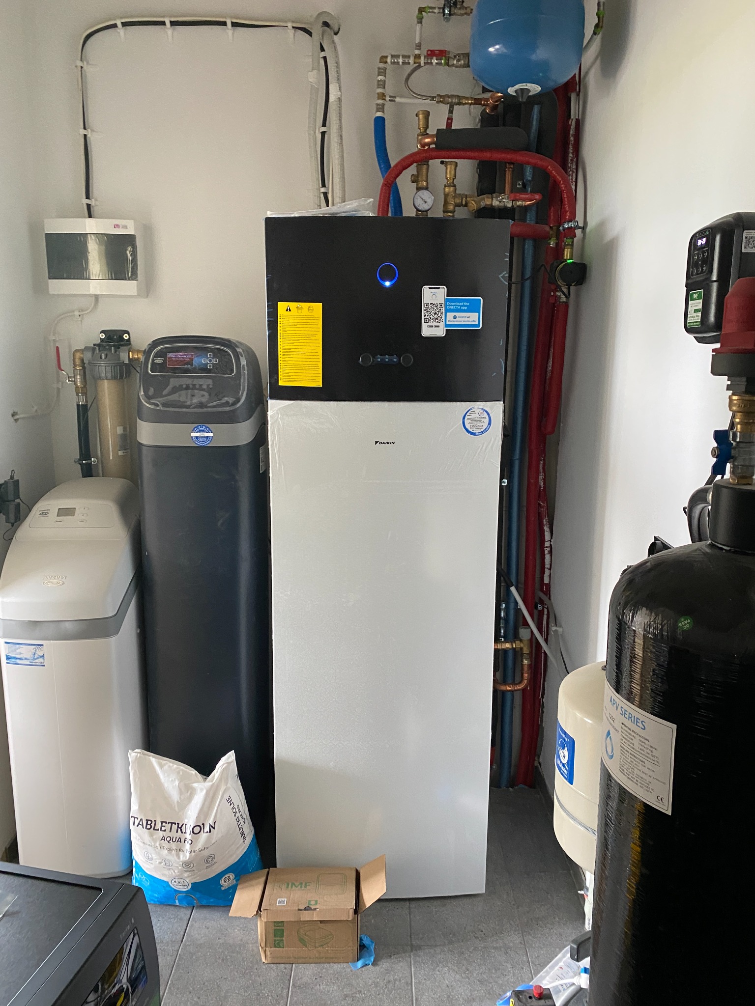 Pompa ciepła Daikin w kotłowni, obok zmiękczacz wody i filtr, widoczne rury instalacji grzewczej, worek z solą i karton. W tle białe ściany i instalacje.
