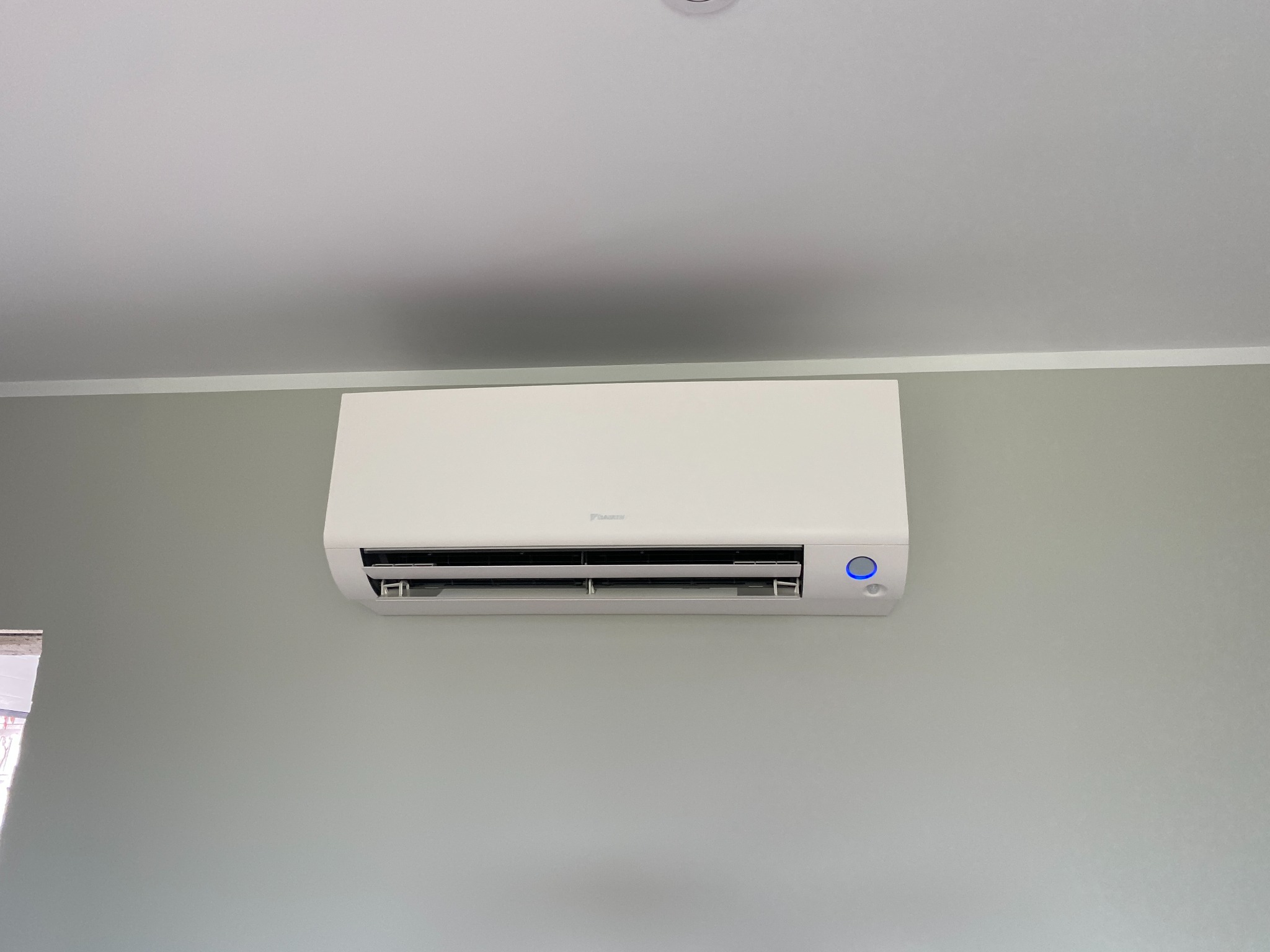 Biała klimatyzacja Daikin zamontowana na szarej ścianie w pomieszczeniu. Widoczny fragment okna po lewej stronie.