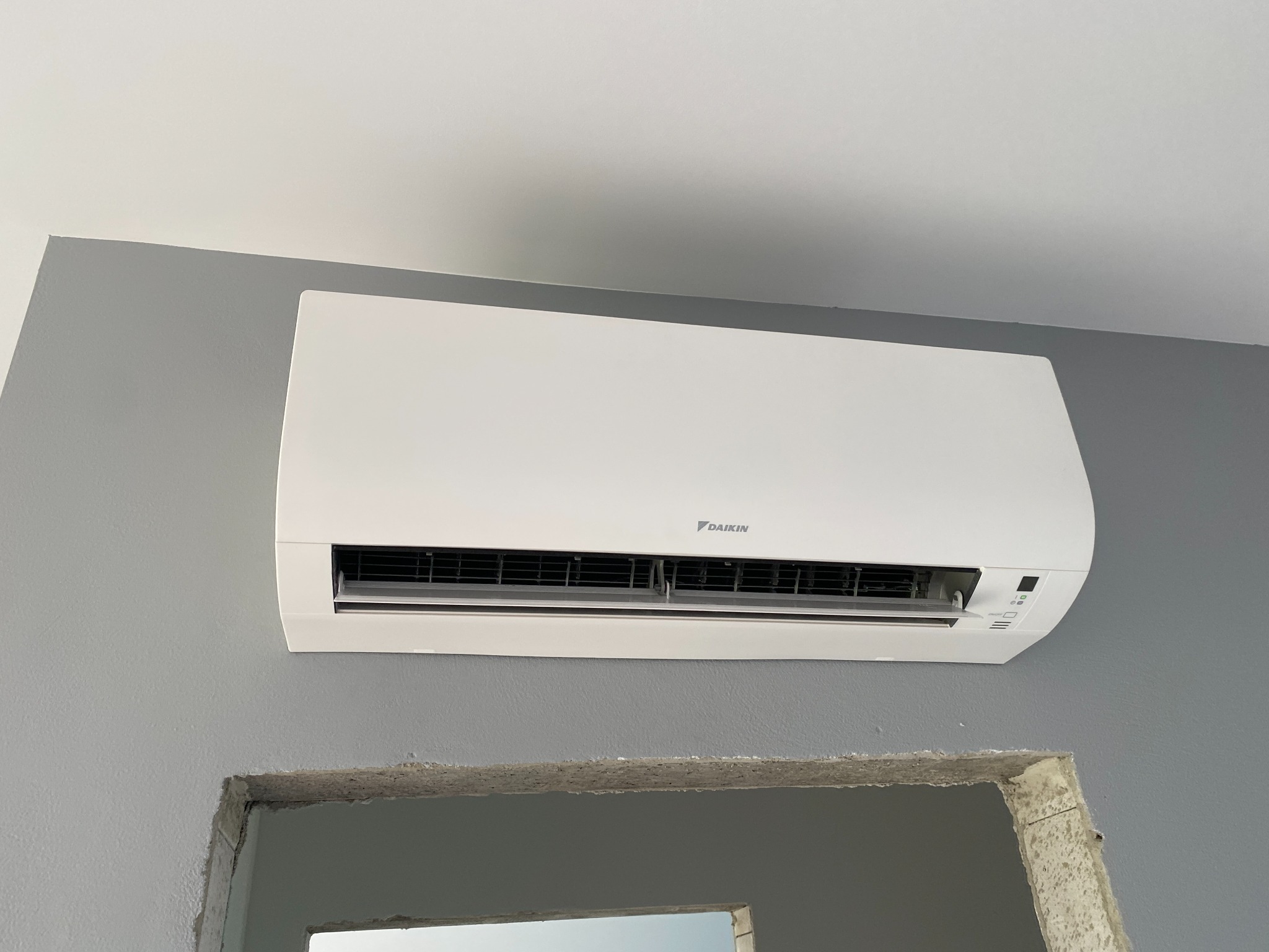 Biały klimatyzator Daikin zamontowany na szarej ścianie nad surowym otworem drzwiowym, widok z dołu.