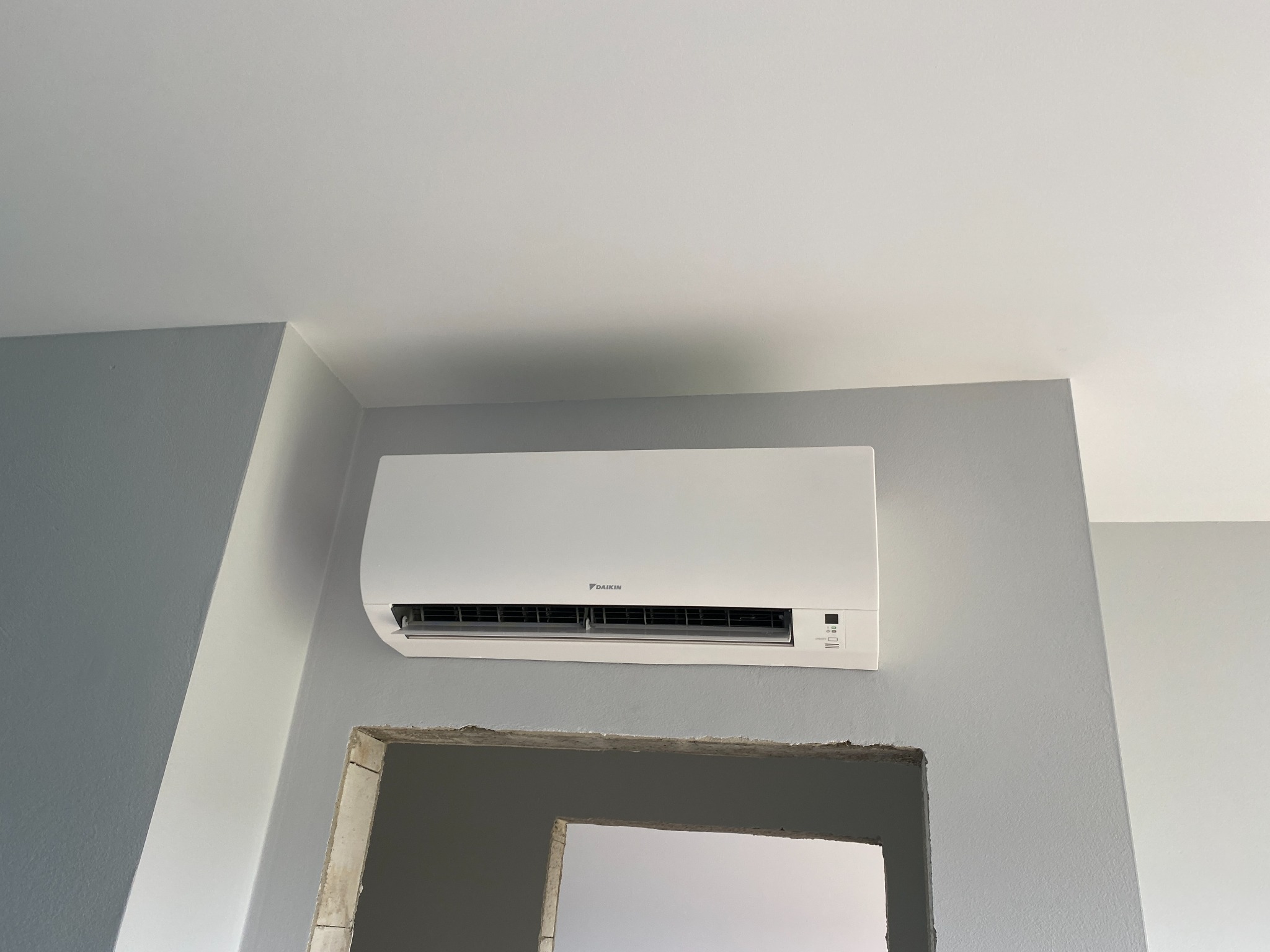 Biała jednostka klimatyzacji Daikin zamontowana na szarej ścianie nad surowym otworem w ścianie. Widok z dołu, na tle białego sufitu.