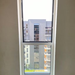 FAMILY GROM SPÓŁKA Z OGRANICZONĄ ODPOWIEDZIALNOŚCIĄ - Nowe białe okno z widokiem na budynek mieszkalny z balkonami, widoczne ślady po montażu na ścianie.