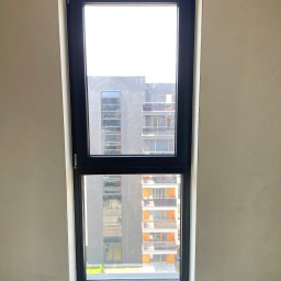 FAMILY GROM SPÓŁKA Z OGRANICZONĄ ODPOWIEDZIALNOŚCIĄ - Wąskie, pionowe okno z czarną ramą, widok na nowoczesny budynek z balkonami, ściana w surowym stanie przed wykończeniem.