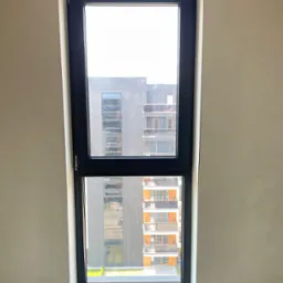 Wąskie, pionowe okno z czarną ramą, widok na nowoczesny budynek z balkonami, ściana w surowym stanie przed wykończeniem.