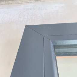 FAMILY GROM SPÓŁKA Z OGRANICZONĄ ODPOWIEDZIALNOŚCIĄ - Szczegółowe zbliżenie na narożnik szarej ramy okiennej z widocznymi śladami niedawnego uszczelniania silikonem i surowym wykończeniem ściany w tle.