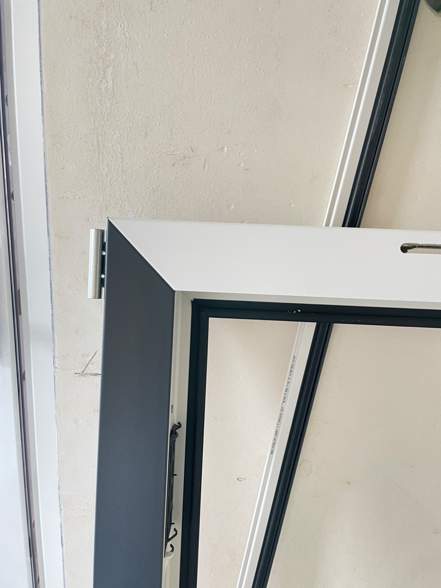 Fragment okna z szarą ramą i metalowym okuciem, widoczne detale mocowania na tle ściany w trakcie montażu lub naprawy.