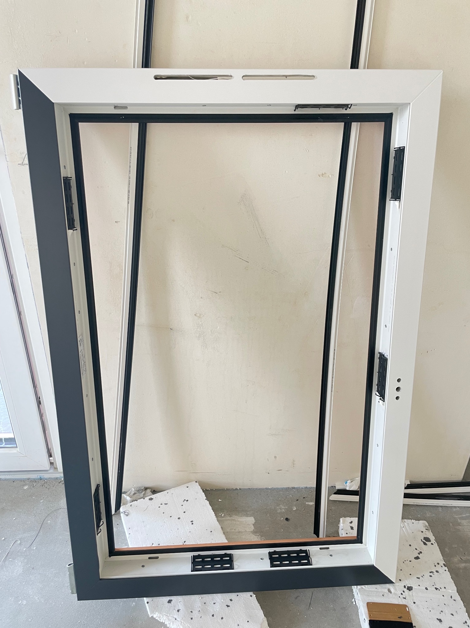 Otwarta rama okienna z widocznymi zawiasami i czarną uszczelką, oparta o ścianę, widoczne fragmenty pianki montażowej na podłodze.