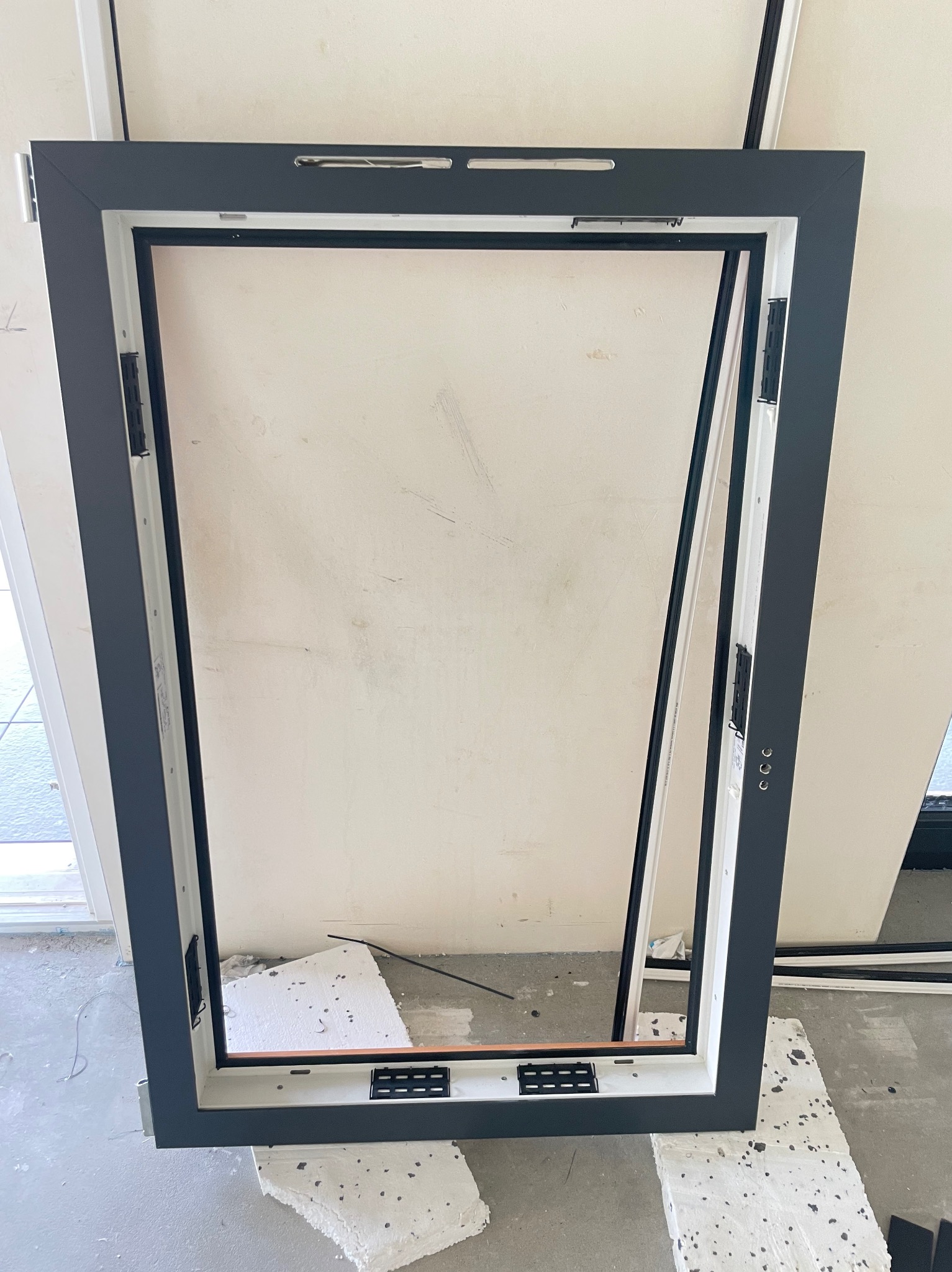 Ciemnoszara rama okienna z widocznymi elementami montażowymi, oparta o białe bloczki styropianowe na szarym, surowym podłożu.