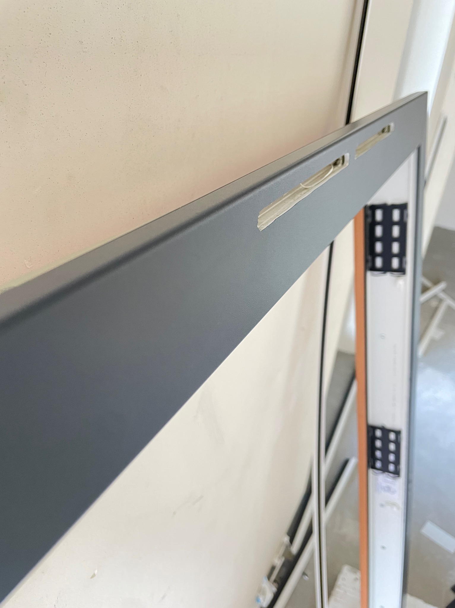 Szary profil okienny z widocznymi otworami montażowymi, zamontowany przy ścianie w trakcie instalacji, z widocznymi elementami mocującymi i częścią ramy okiennej w tle.